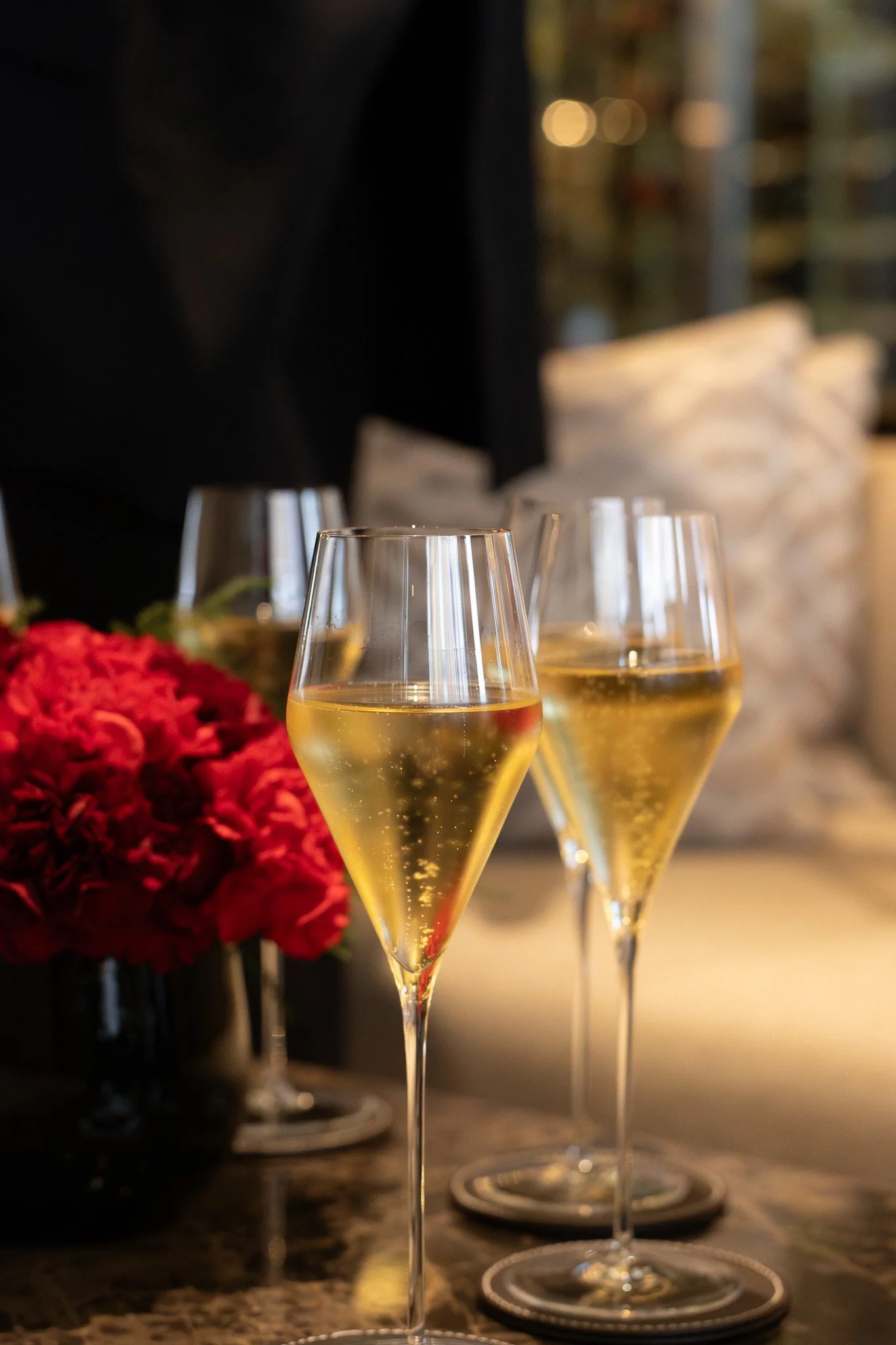 Three glasses of white wine on a table, with a blurred background of a floral arrangement and a dark setting. Petrus.