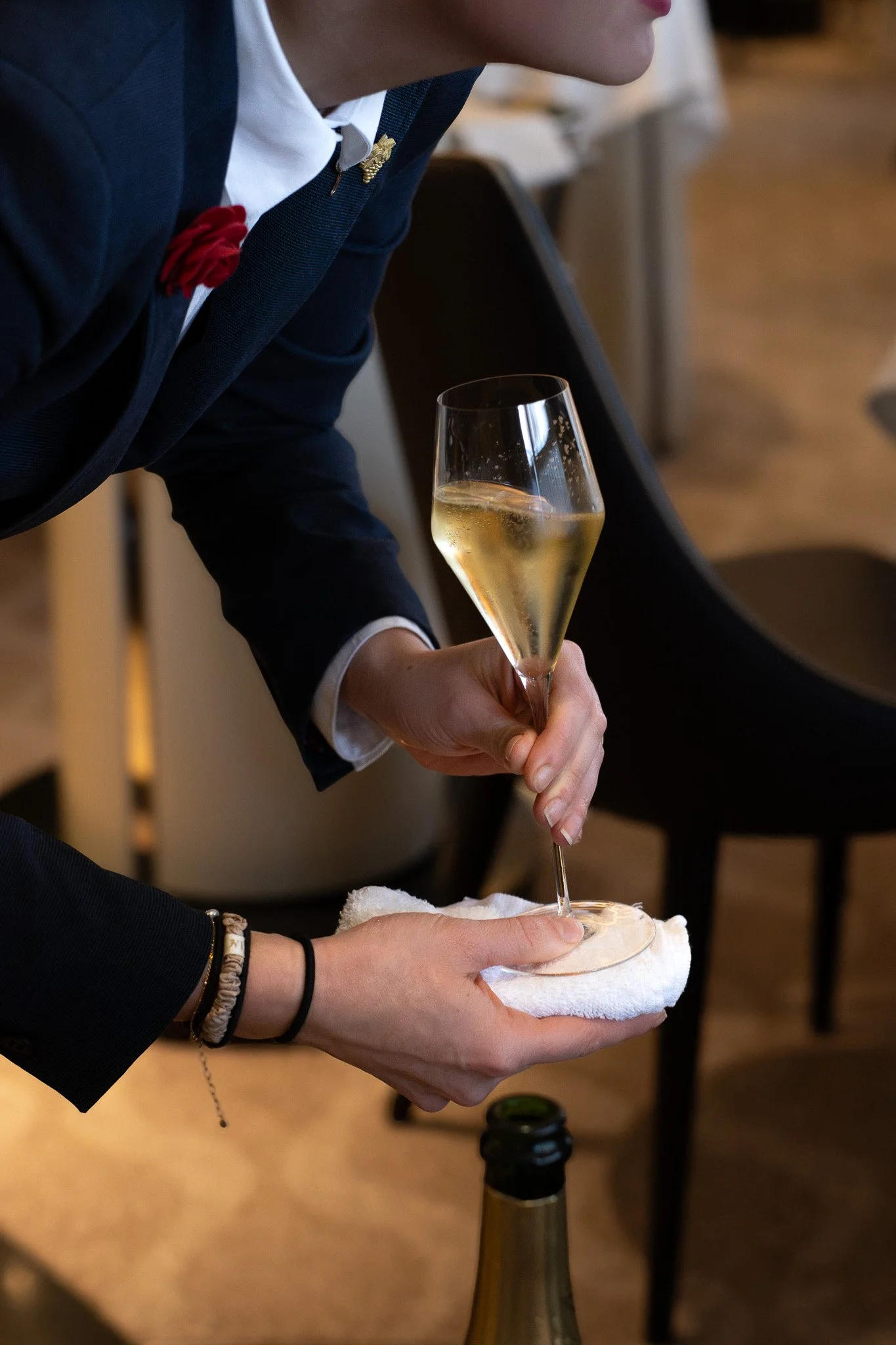 Person holding a champagne glass with white cloth underneath, bottle of champagne on table, wearing dark clothing with bracelet and watch, in indoor setting. Petrus. Server, waitress, sommelier. 