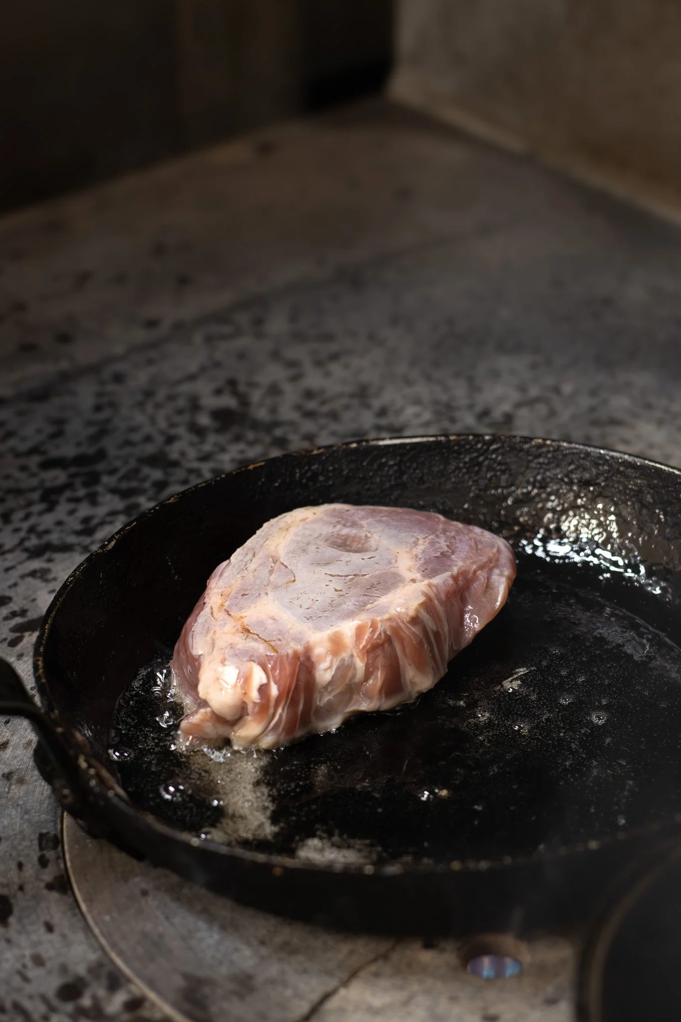 A piece of raw meat being cooked in a black cast iron skillet on a stovetop. The Lamb Inn. 