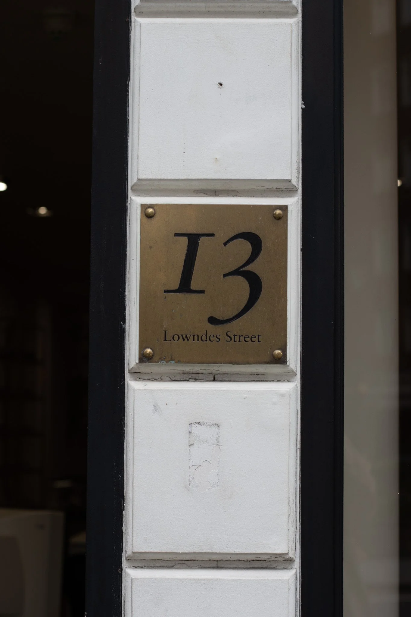 A house number plaque displaying '13 Lowndes Street' mounted on a white post with a black border; the plaque has a gold-colored background with black lettering. Linda Farrow.