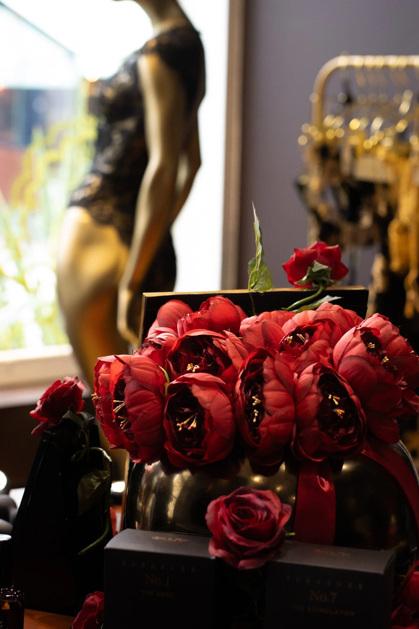 A close-up of a vibrant red rose bouquet in a dark metallic vase in front of a black box with gold text, with a gold decorative piece and blurry artwork, with a window to the left providing natural light. Coco De Mer.