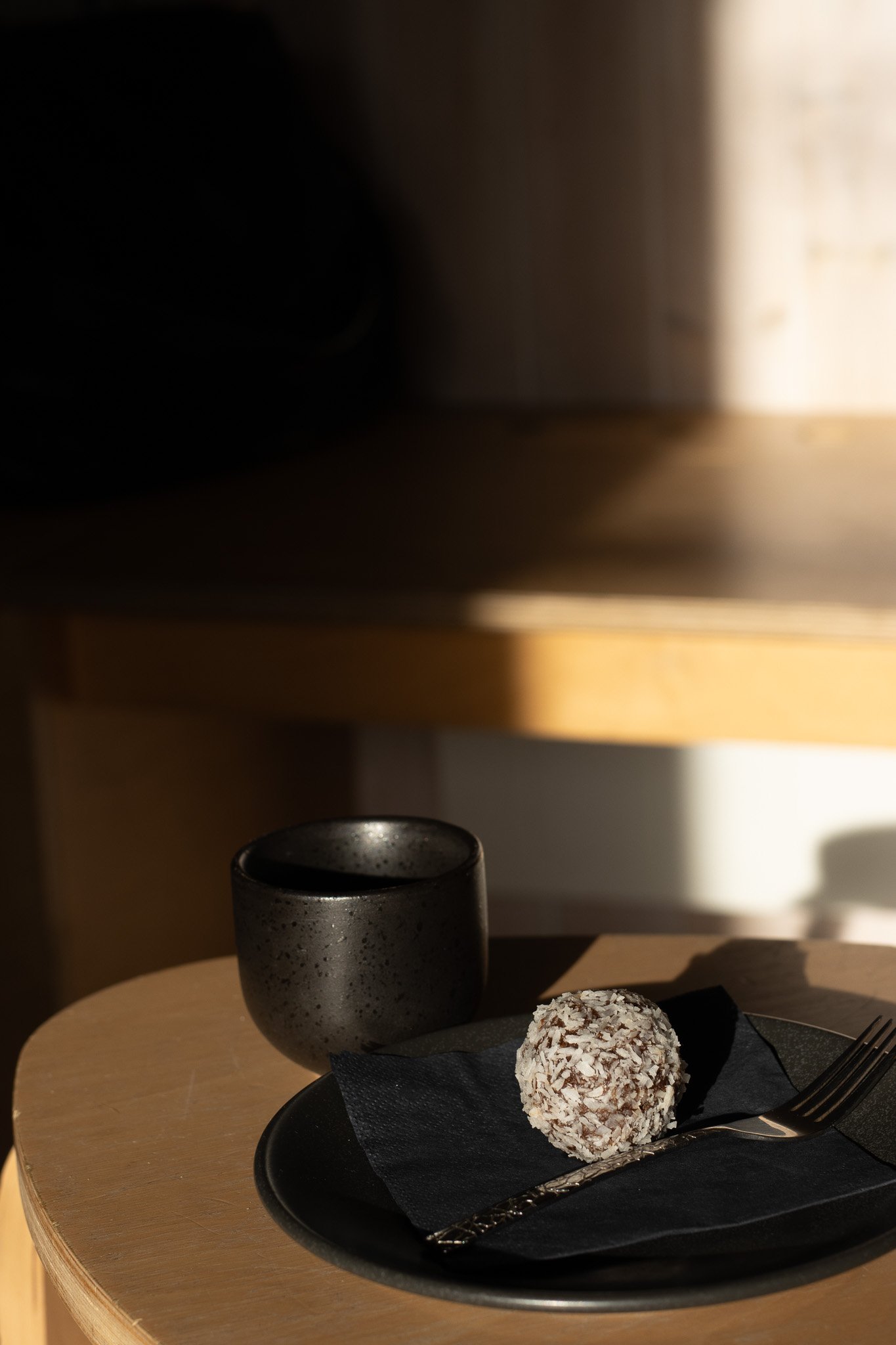 A black plate with a coconut-covered dessert, a fork, and a napkin, on a light wooden table, with a black cup and a ceramic container in the background. The Scandinavian Coffee Pod.