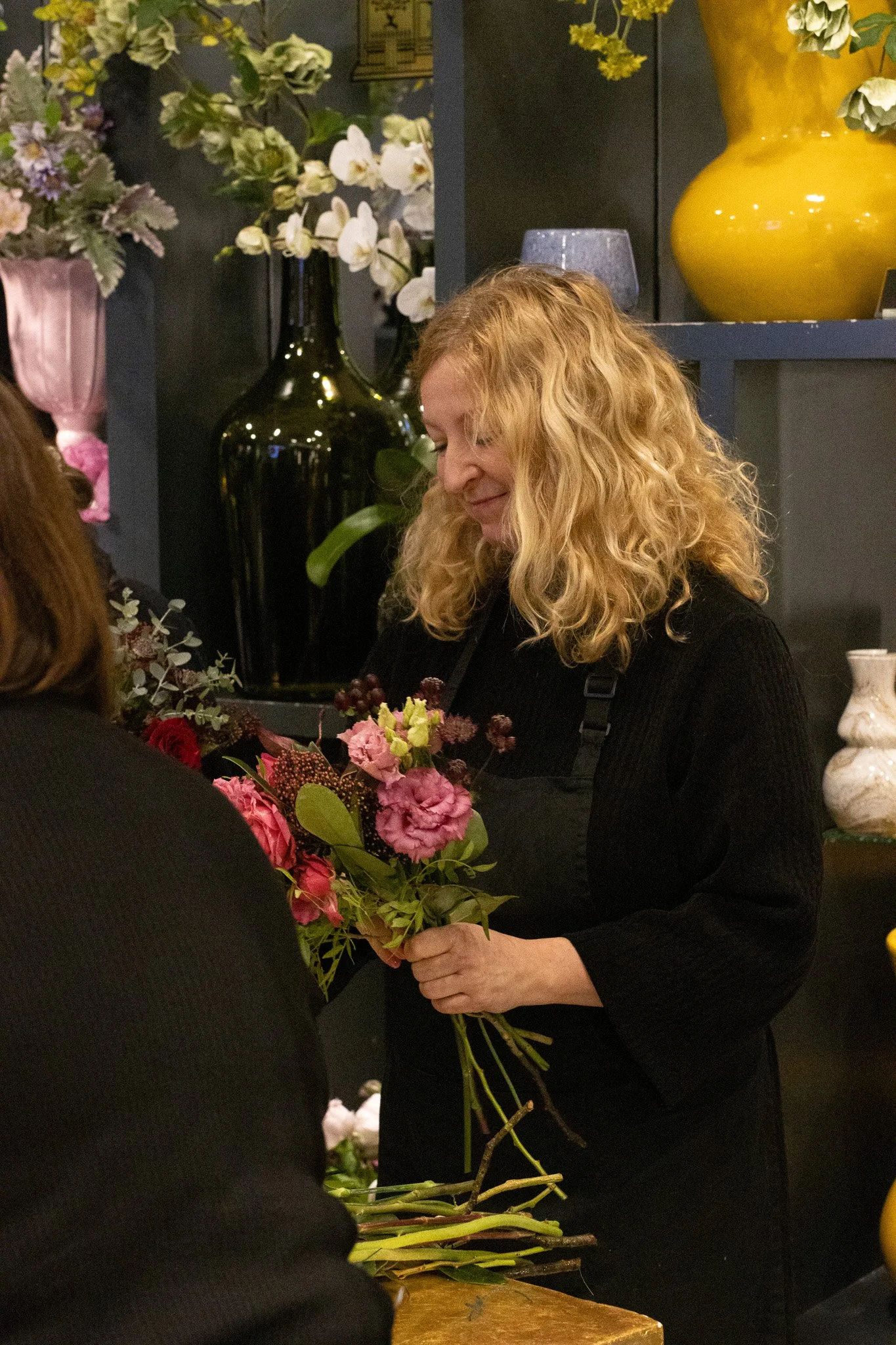 A woman with curly blonde hair, wearing a black top, is holding a bouquet of pink, purple, and green flowers. She is smiling softly and looking down at the flowers. Behind her, there are large decorative vases with white and yellow flowers, and a sec