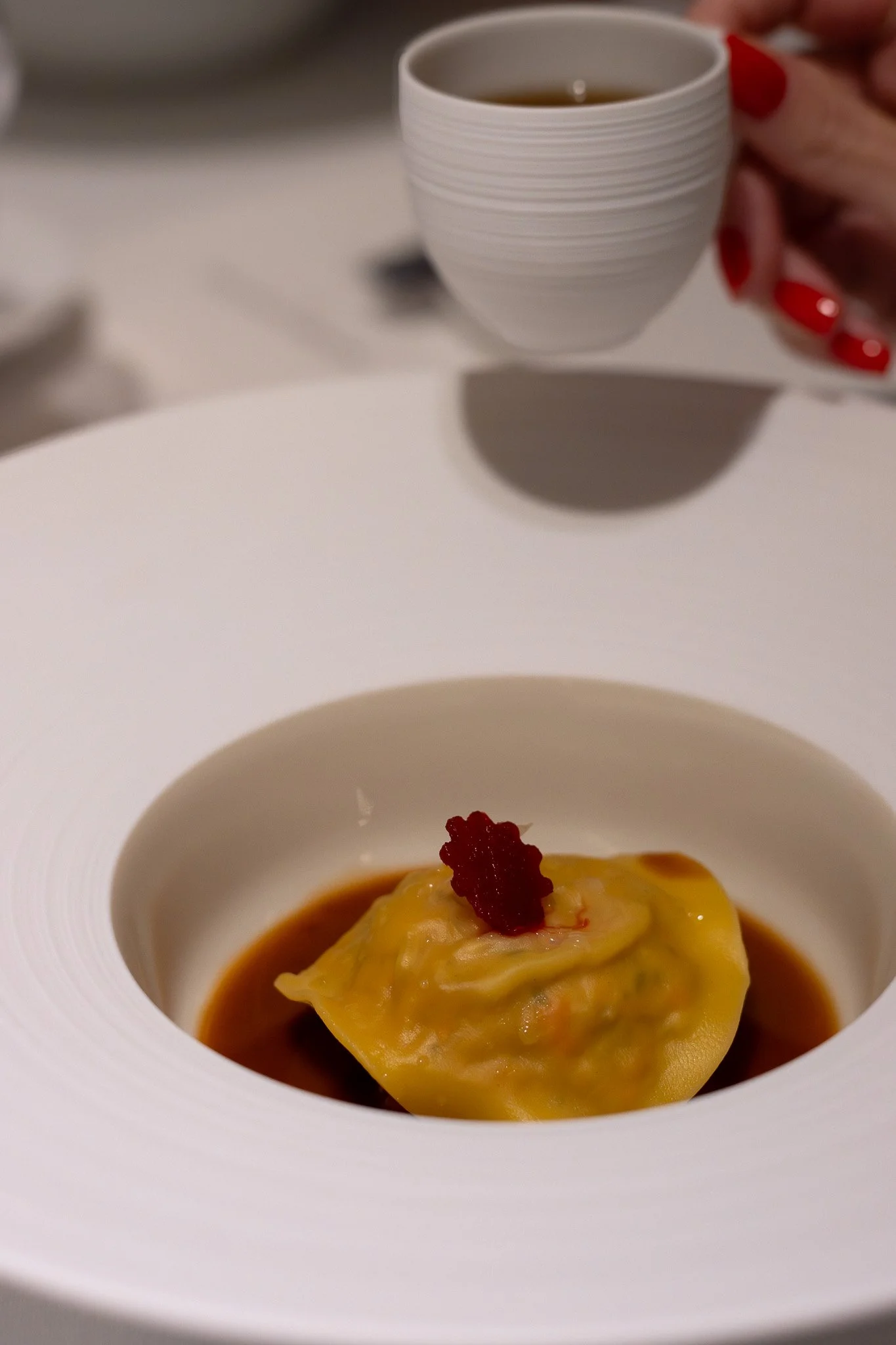 A close-up of a white bowl with a piece of ravioli topped with a red garnish, surrounded by dark sauce. Petrus.