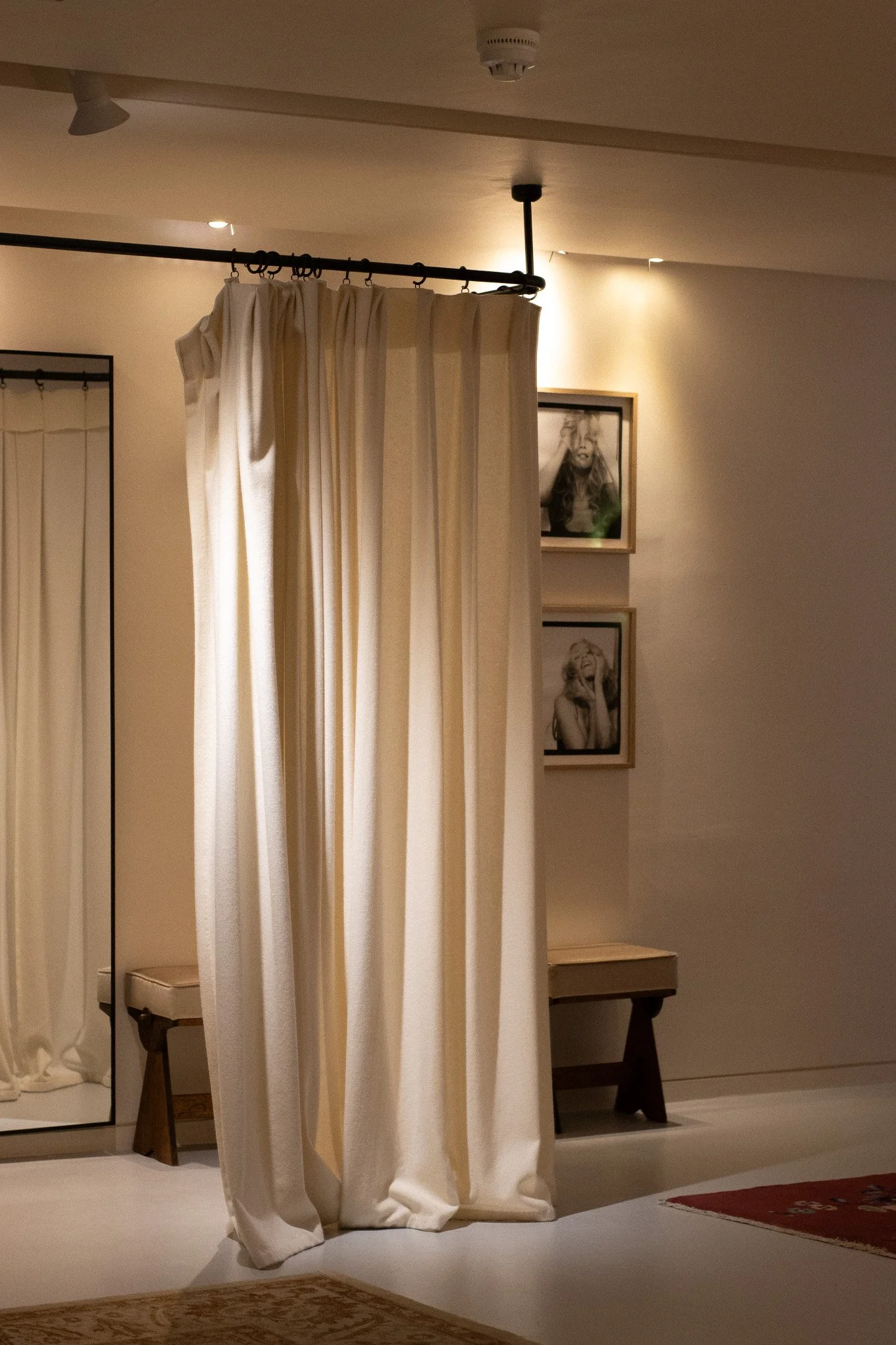 A room with white curtains hanging from a black curtain rod, a framed black-and-white portrait of a woman with long hair and a hand on her head, a small wooden side table, and a patterned area rug on the floor. Realisation Par.