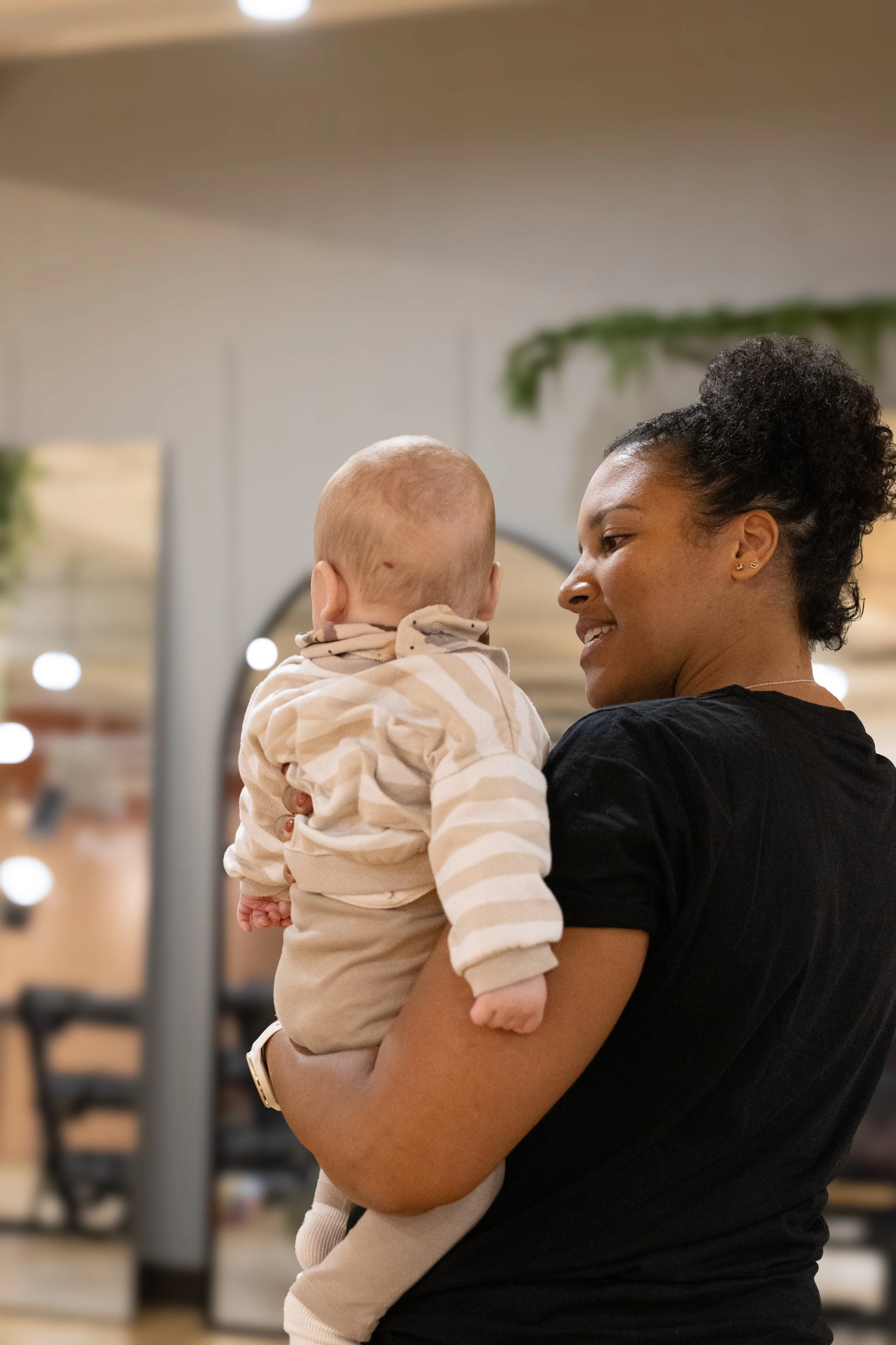 A woman holding a baby while looking at each other, in an indoor setting. The Studio Cheltenham, Reformer Pilates. 