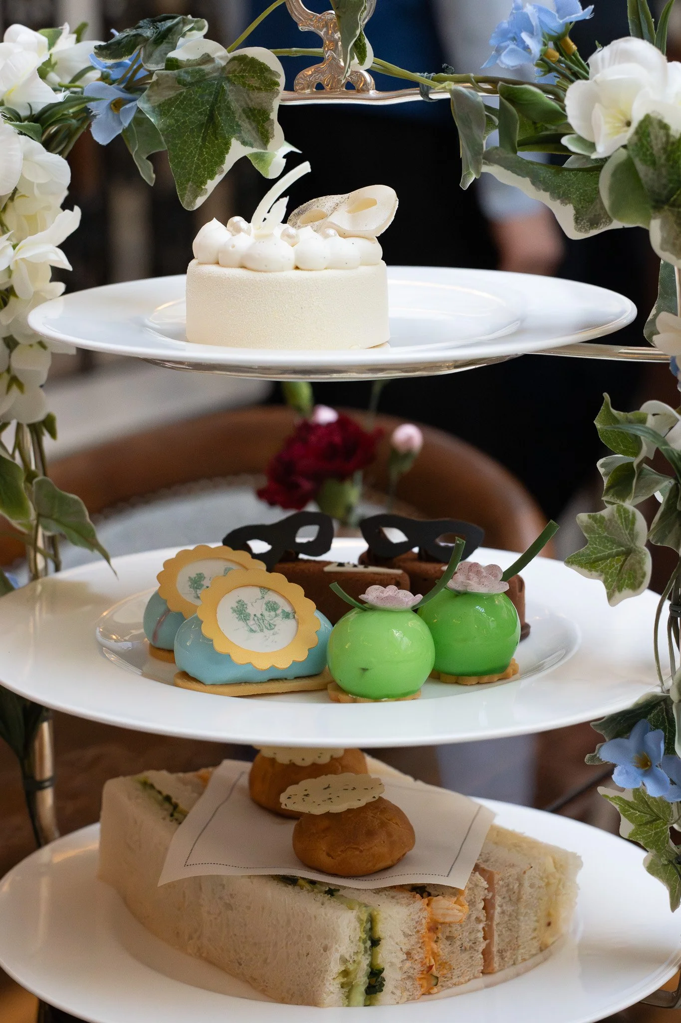  Bridgerton Afternoon Tea, with assorted desserts, including a white cake on the top, decorated with white frosting dollops and a white chocolate leaf, adorned with a floral arrangement of green leaves, white and blue flowers.  The Lanesborough.