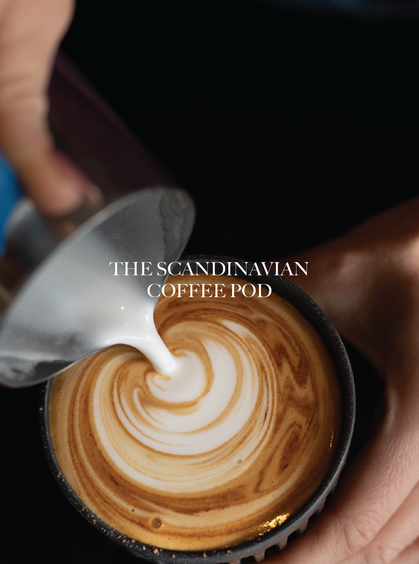 Close-up of a hand pouring steamed milk into a cup of coffee, creating a latte art pattern with a cream swirl design. The Scandinavian Coffee Pod.