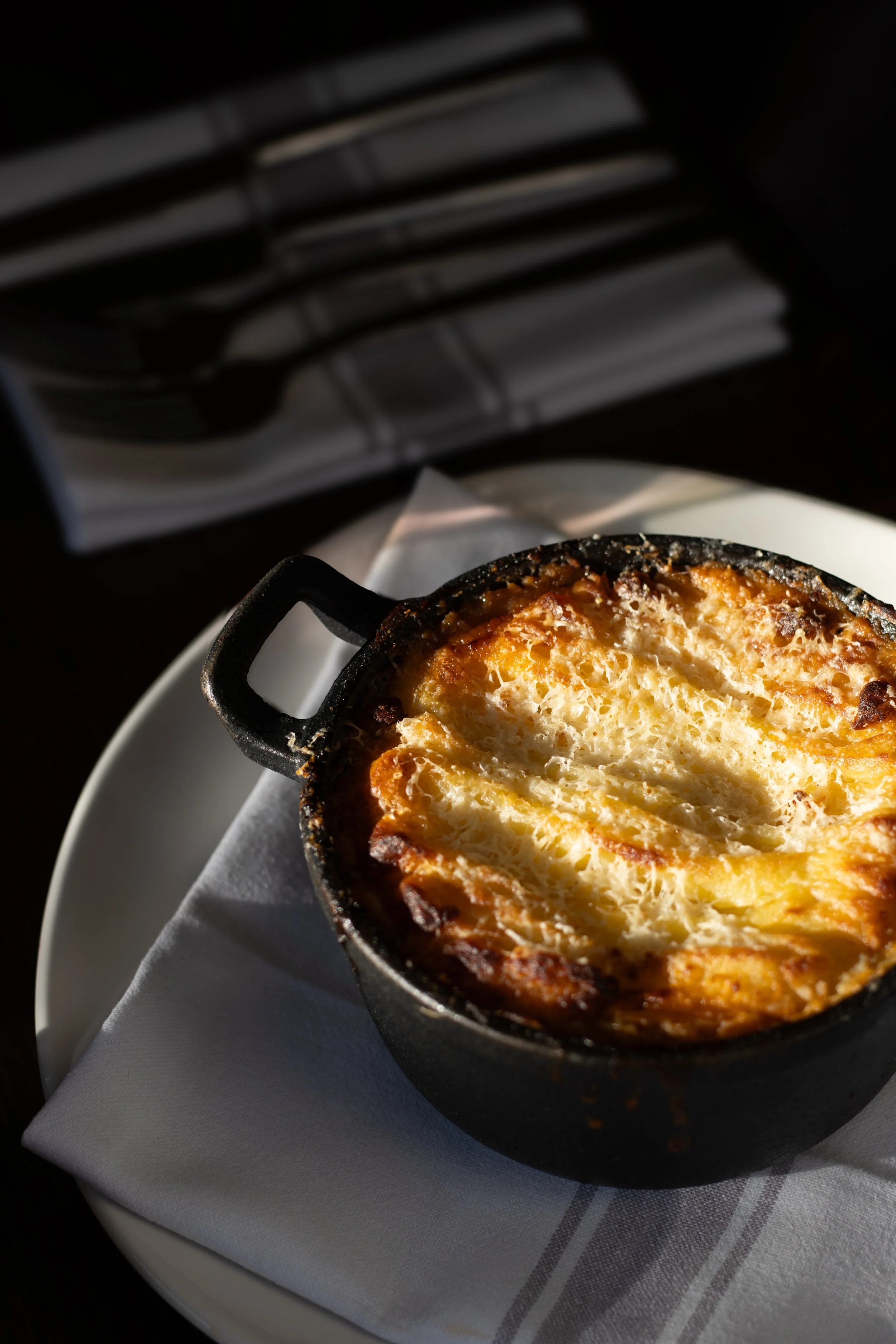A small black cast-iron skillet containing a baked dish with a golden, crispy top, possibly a gratin or casserole, on a white plate with a cloth napkin underneath, with sunlight casting shadows. The Lamb Inn. 