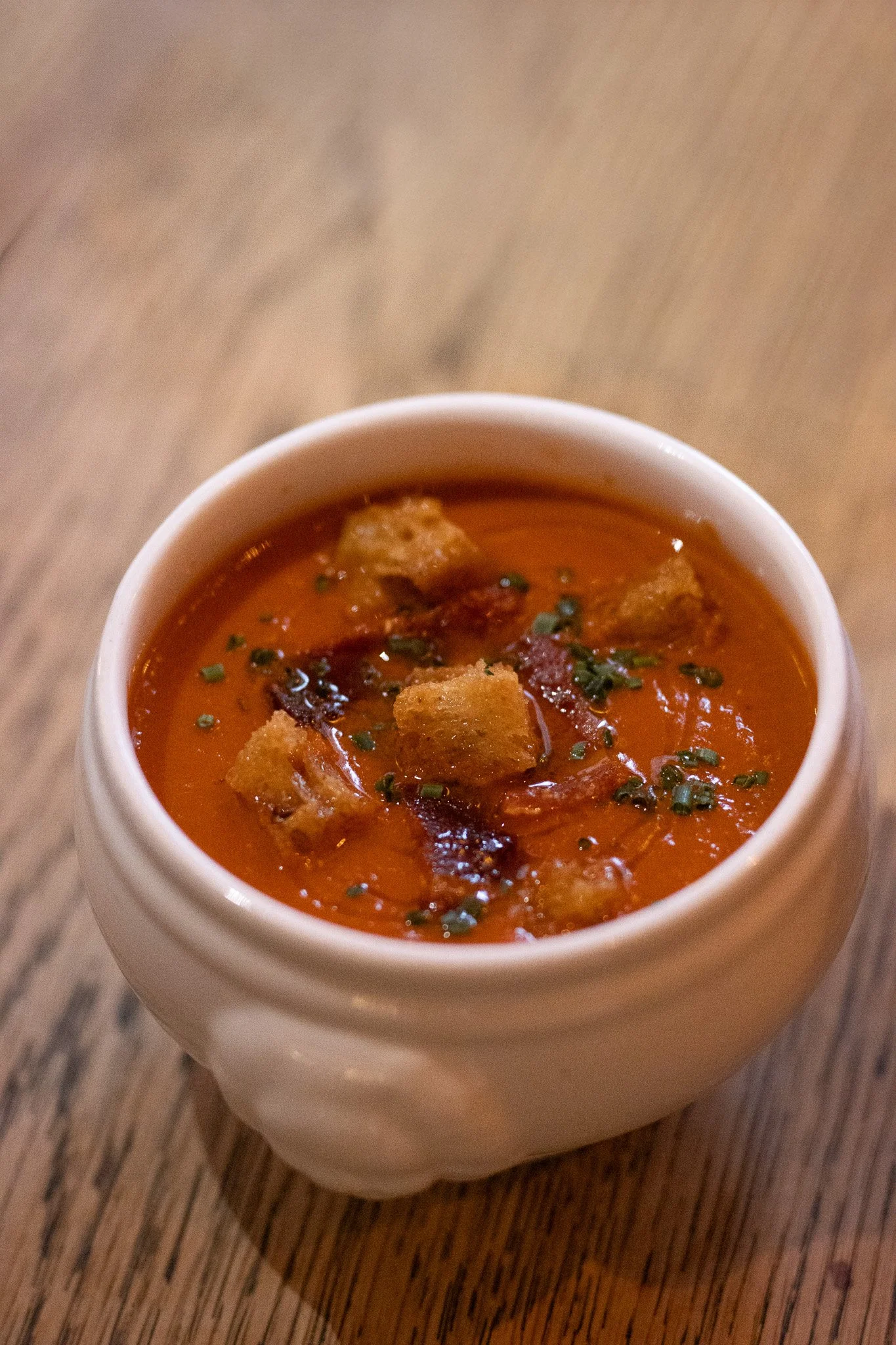 A white ceramic cup filled with tomato-based soup or stew garnished with black pepper and chopped herbs, placed on a wooden table. Alfred Tennyson.