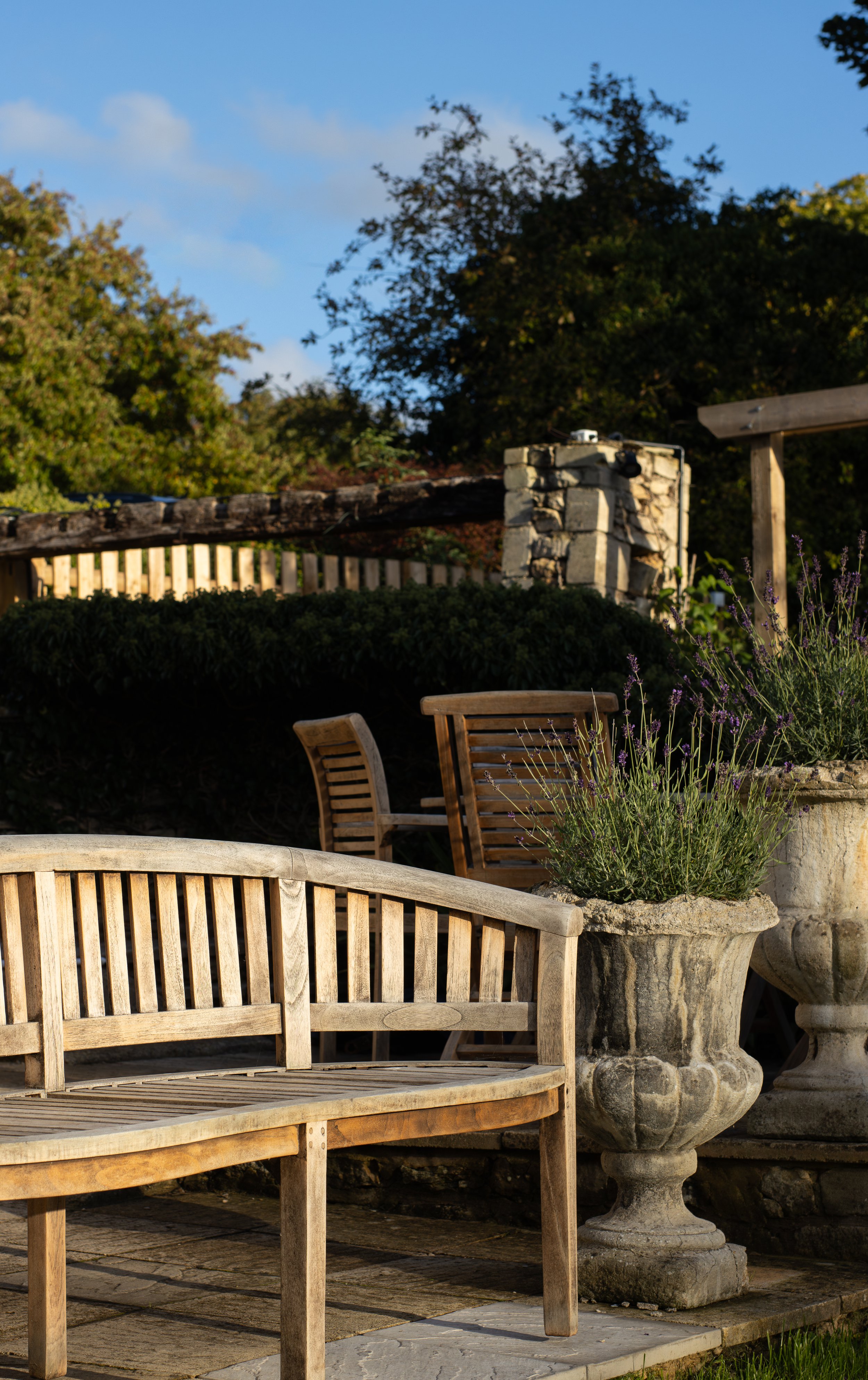 Outdoor garden scene with a wooden bench in the foreground, a large stone planter with purple flowers, a stone pillar, and a wooden fence with lush green trees and blue sky in the background. The Lamb Inn. 