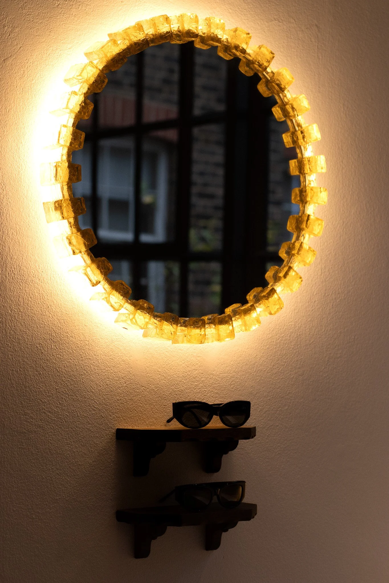 A decorative mirror with a yellow gemstone border hanging on a wall, illuminated by a light behind it. Below the mirror, a small wall shelf holds a pair of black sunglasses with dark lenses. Realisation Par.
