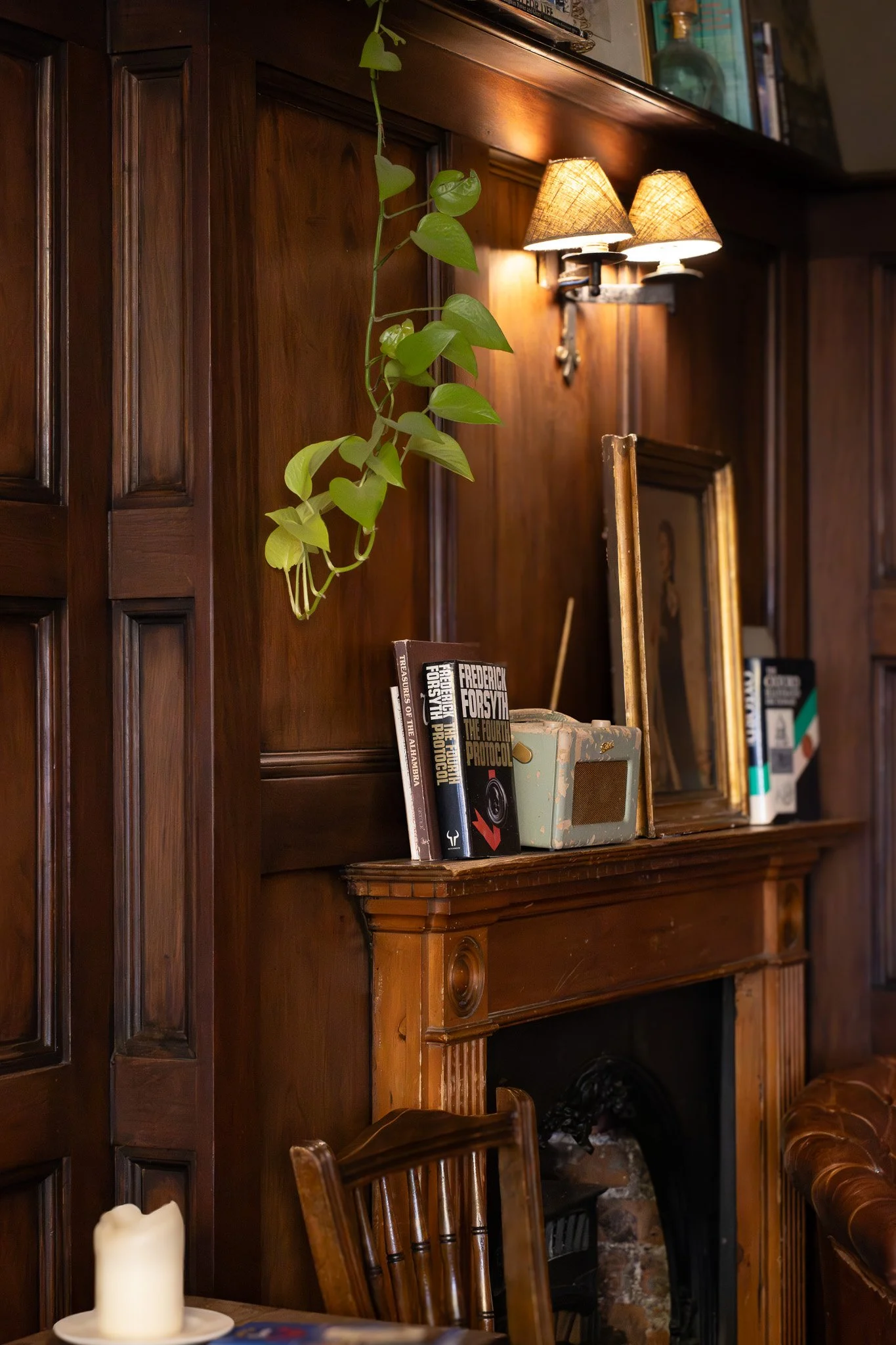 A wooden fireplace mantle and wall, with a green leafy hanging plant, a small bookshelf with books, a mirror, and a wall sconce with two lamps. Part of a table with a candle and a chair are visible in the foreground.  The Wilton Arms.