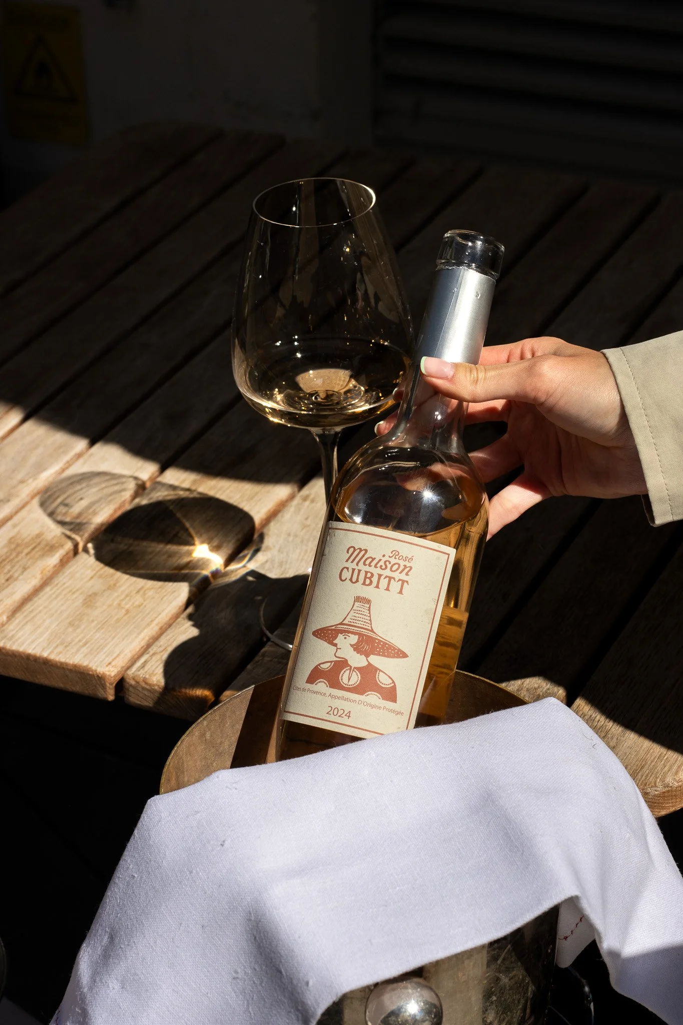 A hand holding a bottle of Maison Cubitt rosé wine next to a wine glass with some wine, on a wooden table outdoors, with sunlight casting shadows. GetAGrip Apparel. 