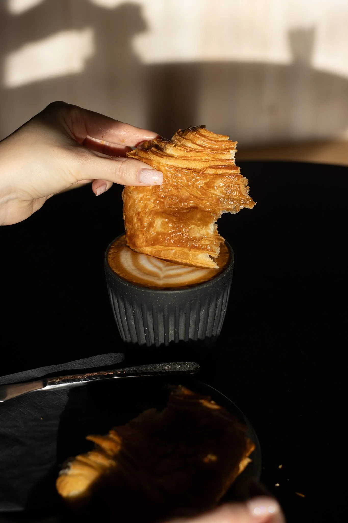 A hand holding a piece of flaky, crispy pastry over a cup of coffee with latte art. The background is blurred. The Scandinavian Coffee Pod.