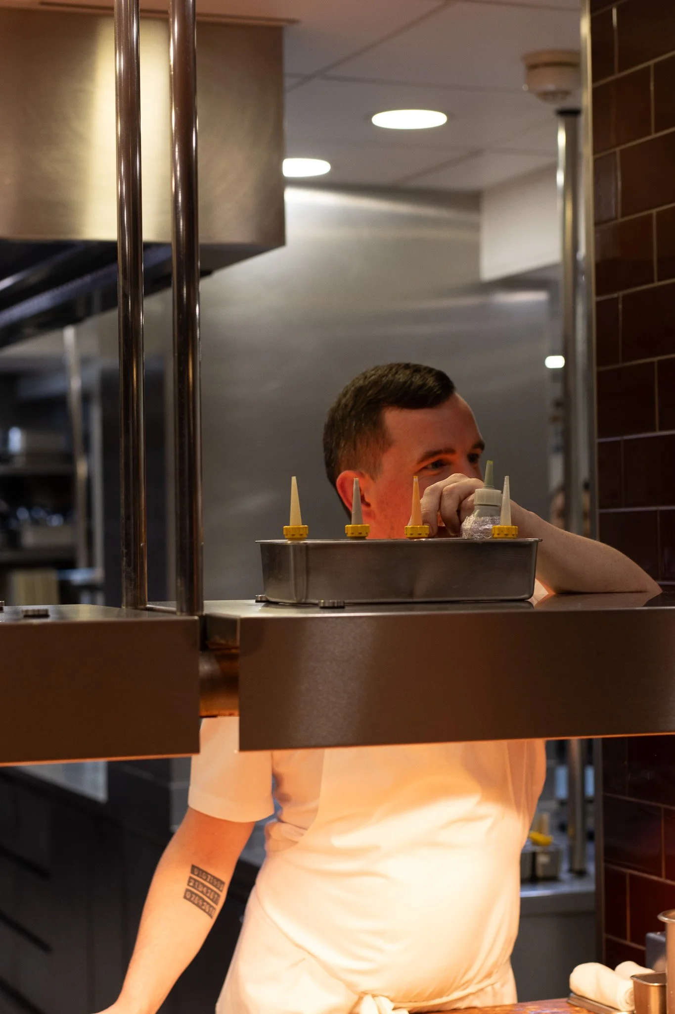A person with short hair and a tattoo on their left arm, wearing a white shirt, is working behind a counter in a restaurant or kitchen. The background shows a commercial kitchen setting with stainless steel. ORSON VERGNAUD. Petrus.