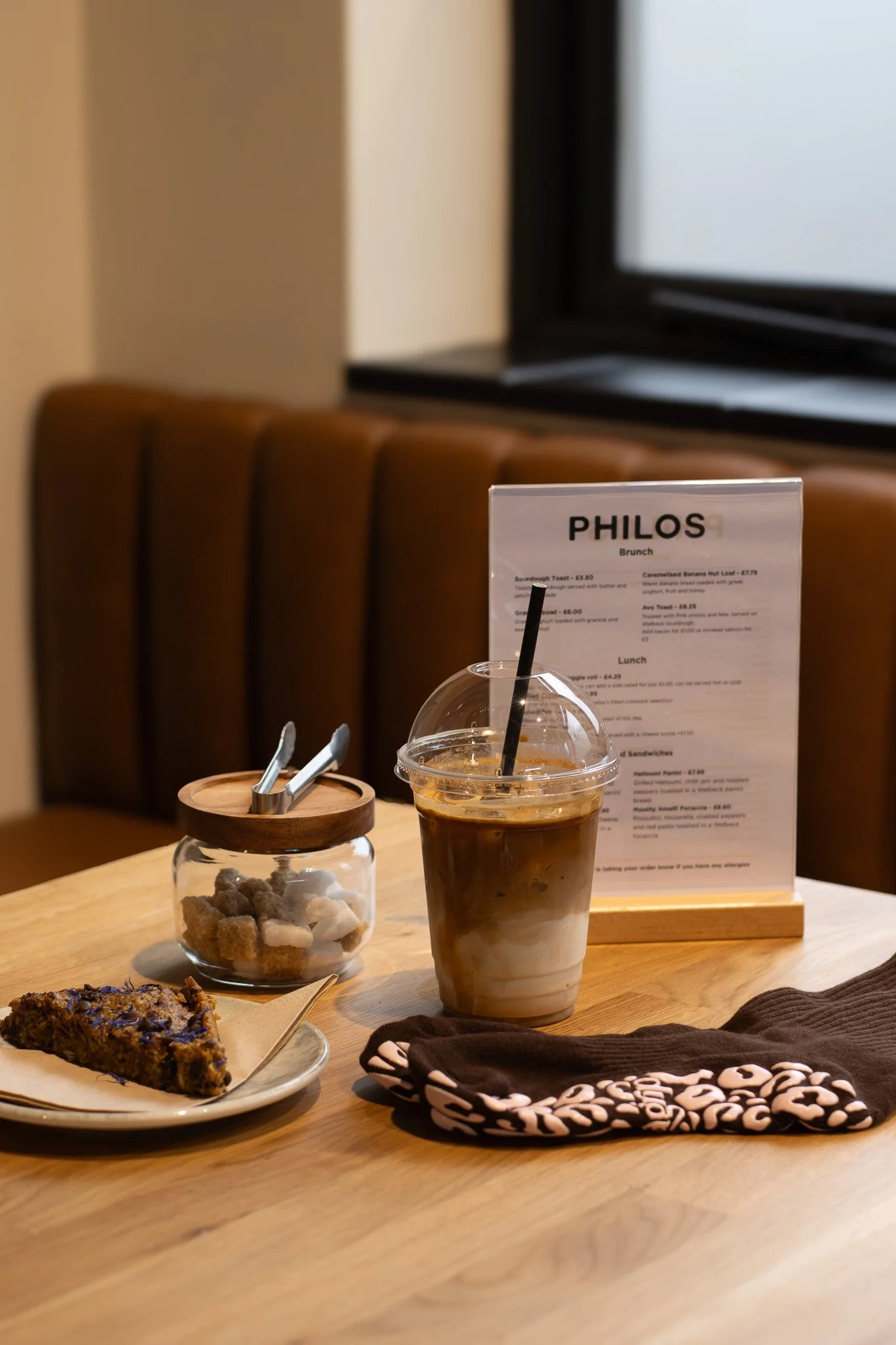 A table with a caramel frappuccino in a clear plastic cup with whipped cream on top, a small glass jar with sugar cubes, a slice of chocolate cake, a printed menu, and a folded cloth napkin with a black and white pattern. In the background, there is 