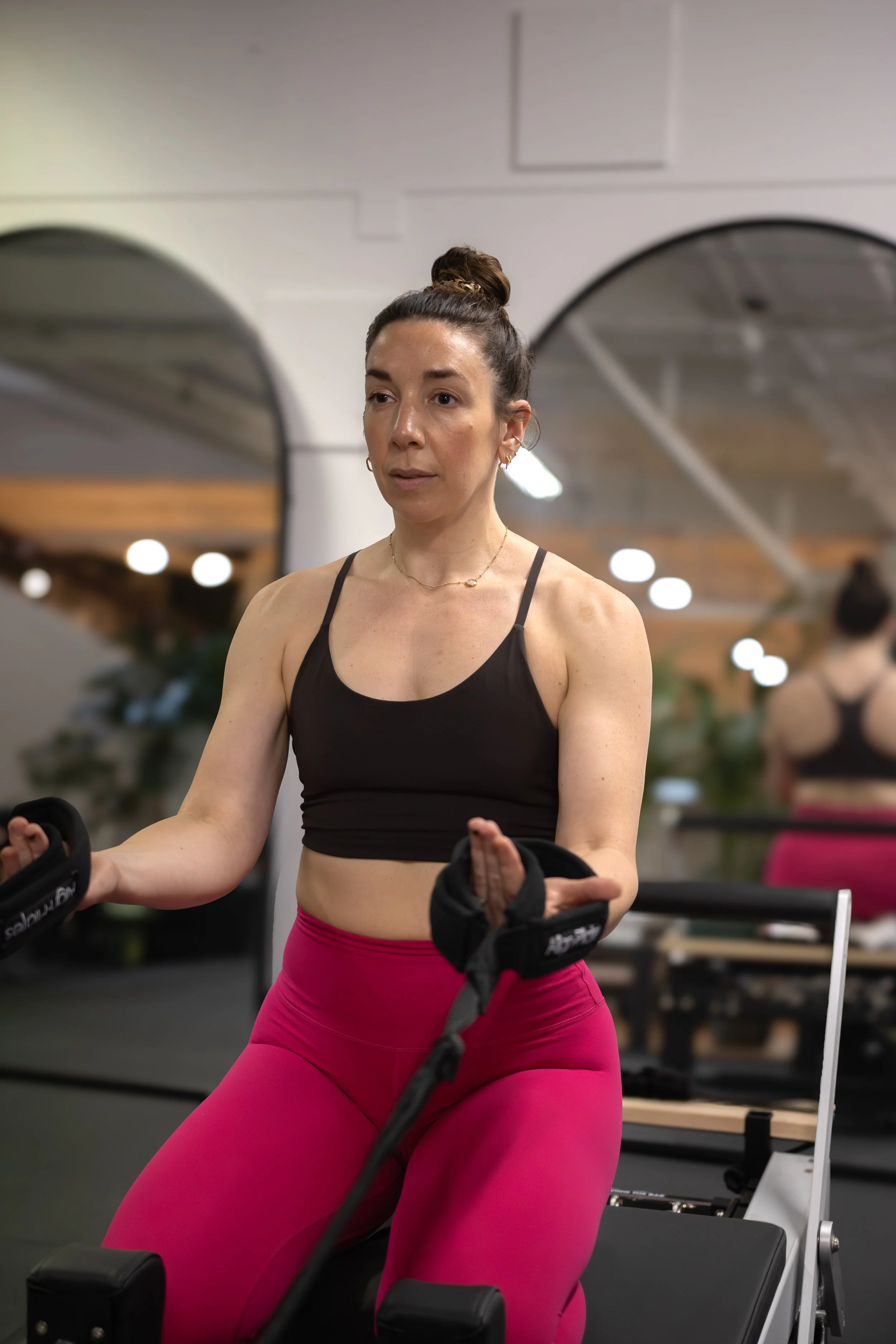 A woman with dark hair tied in a bun, wearing a black sports bra and bright pink leggings, is exercising on a rowing machine in a gym. The background shows mirrors and gym lighting. The Studio Cheltenham, Reformer Pilates. 