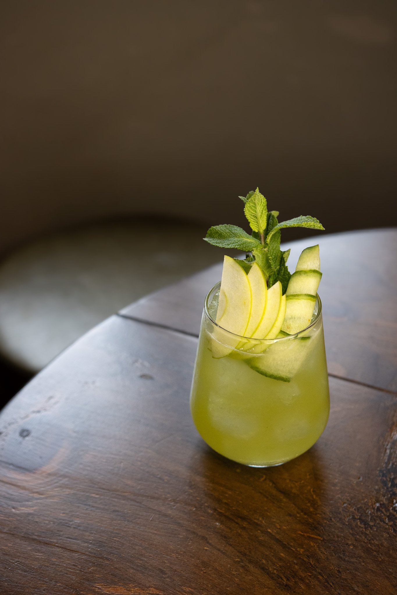 A glass of yellow-green beverage garnished with apple slices and mint leaves, placed on a wooden table. The Lamb Inn. Cocktails. Mixology.