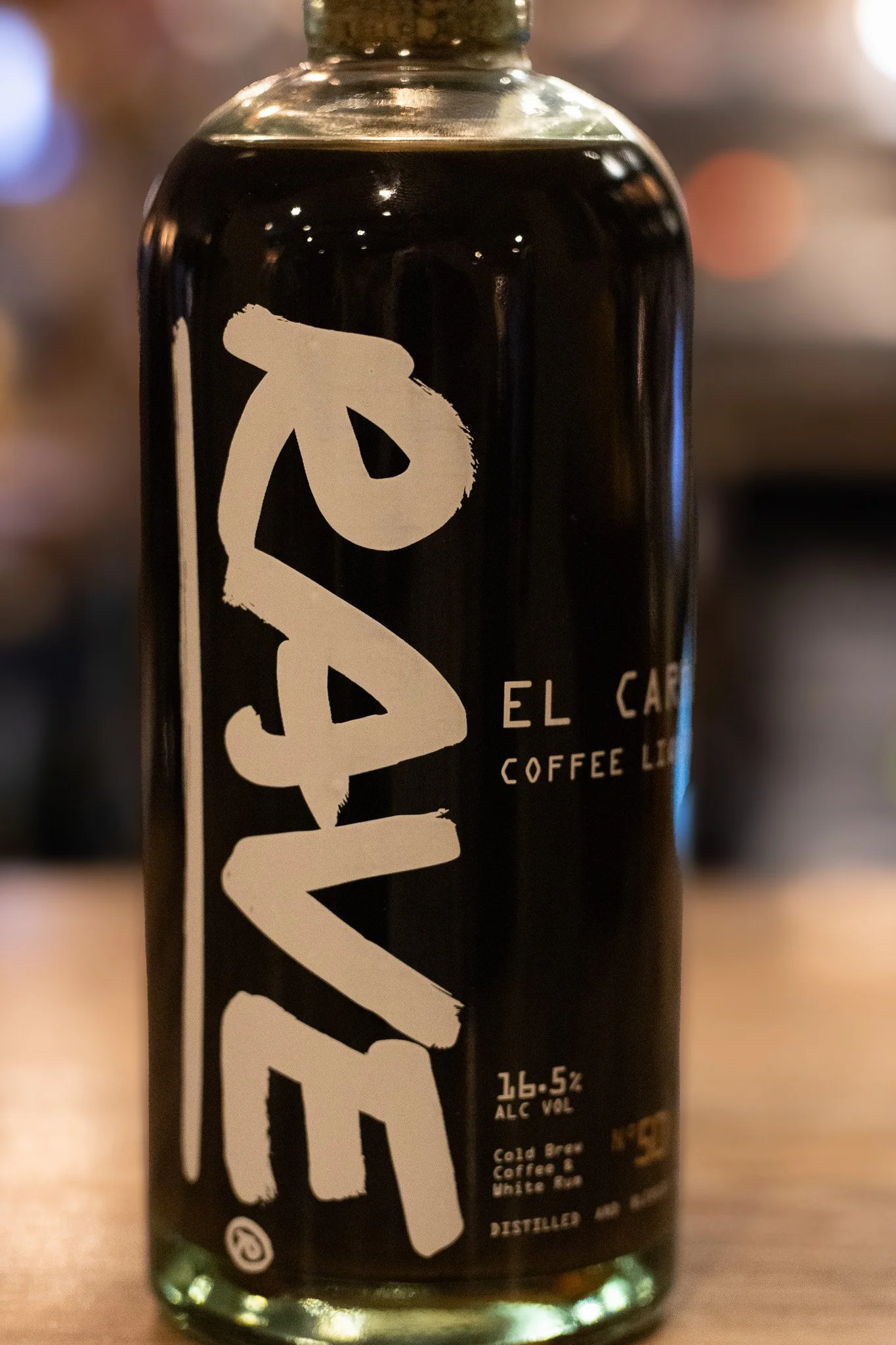 Close-up of a dark bottle with a white label that has a large, stylized text and smaller text, including alcohol content and beverage type, likely a craft beer or alcoholic beverage label. The Lamb Inn. 