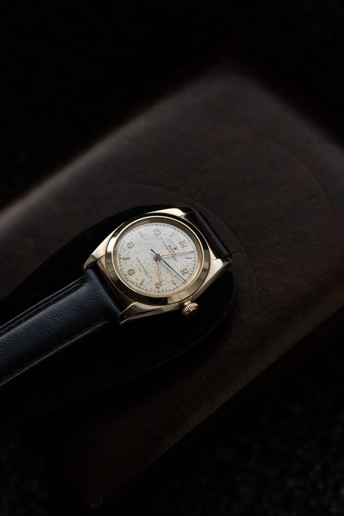 A wristwatch with a light-colored face, gold case, and black leather strap resting on a dark surface. Aurmont. Watches.