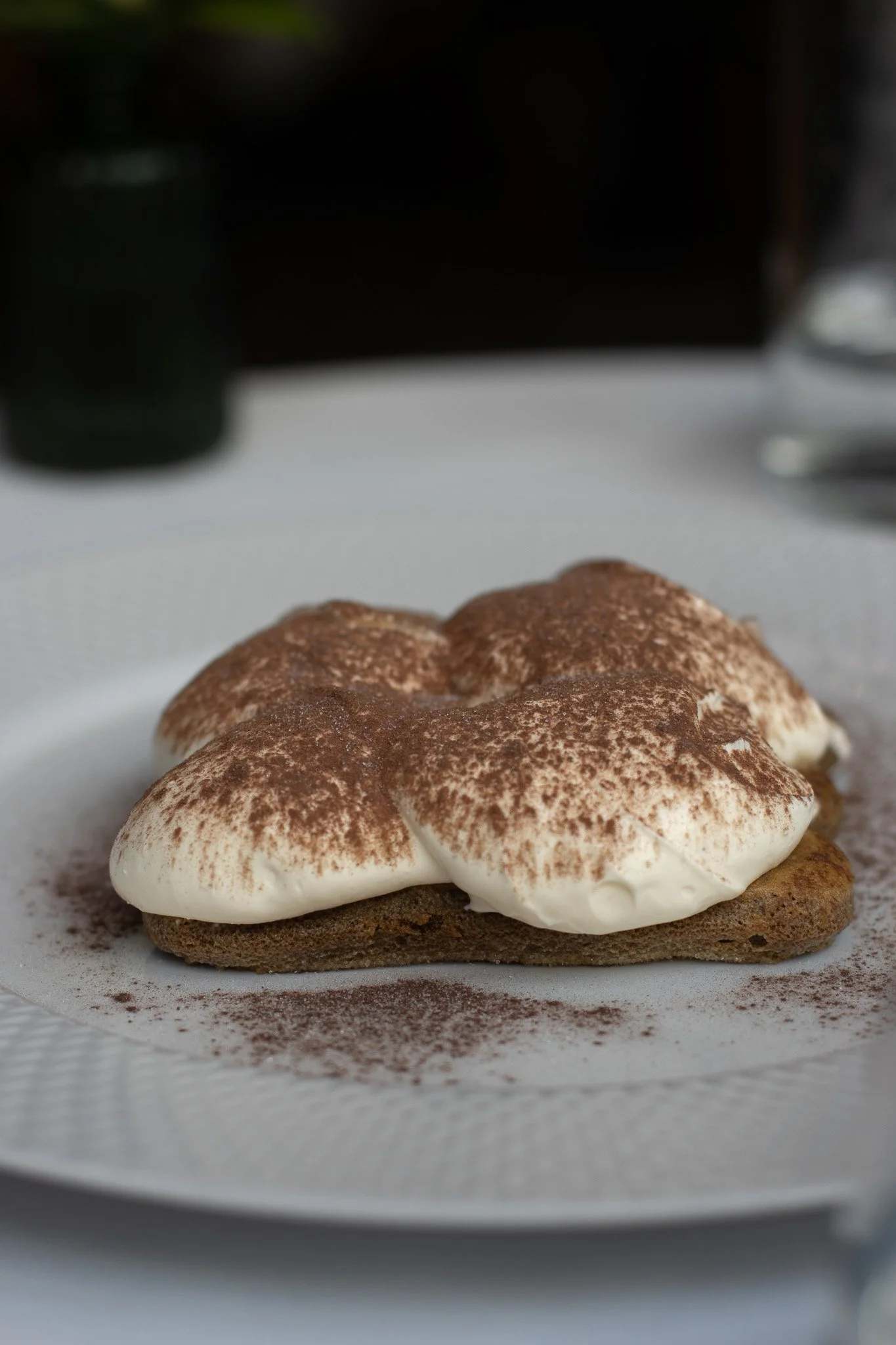 Close-up of a dessert with a cookie base, topped with whipped cream and cocoa powder, on white parchment paper. The Hari, Il Pampero.