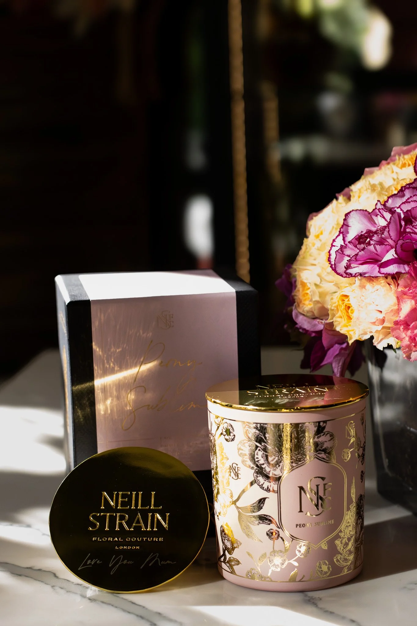 A collection of luxury products including a pink floral scented candle, a black and gold container with the name Neill Strain, and a pink box with gold accents. Neill Strain, Harrods, Motcomb Street.