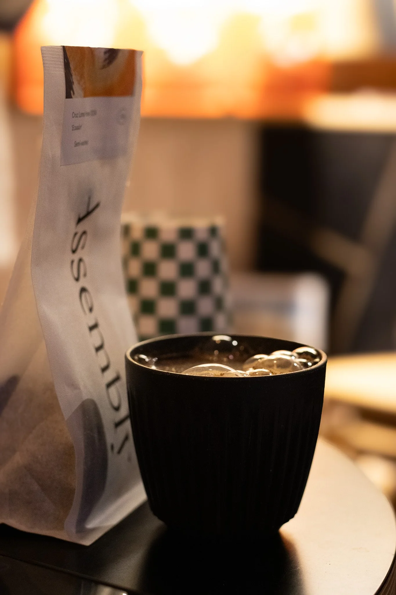 A paper bag with a coffee shop logo, a black cup with coffee and ice, and a checkered cup in the background, likely in a café setting. The Scandinavian Coffee Pod.