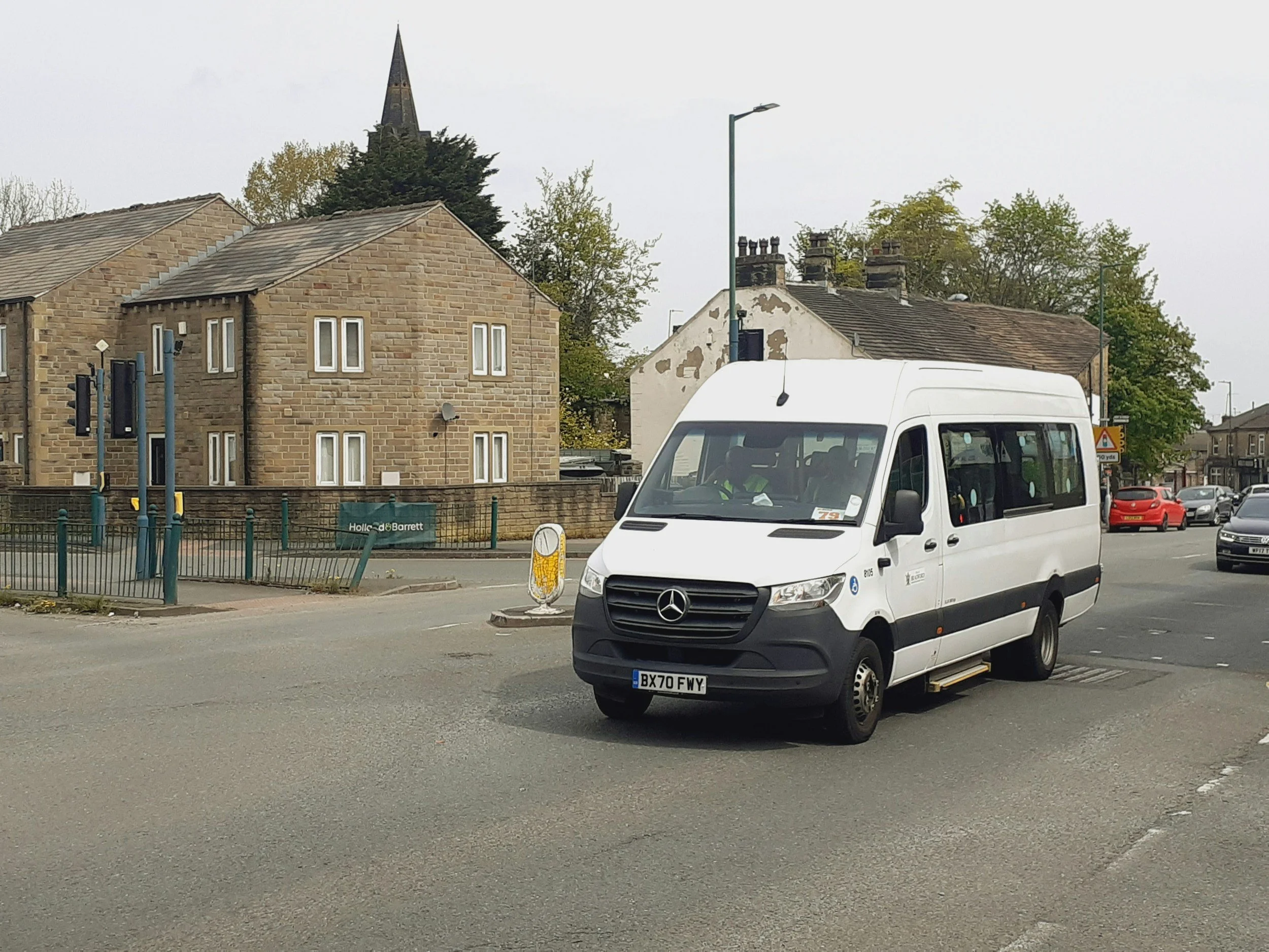 A white Mercedes-Benz minibus stopped at a street intersection in a small town with residential buildings and a church in the background.