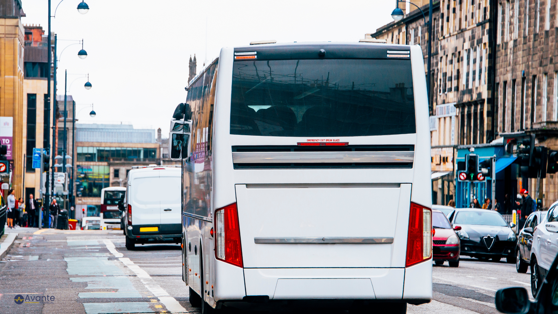 White bus stopped at a city intersection with buildings, cars, and pedestrians around.