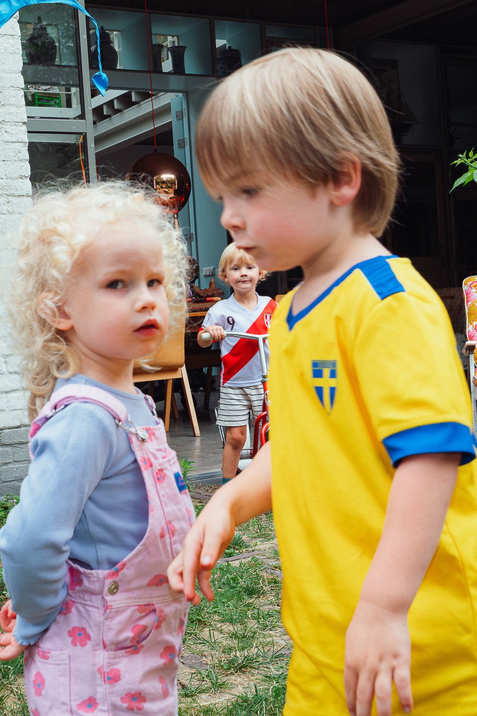 Twee kinderen, een meisje met blond, gekruld haar en een jongen met lichtbruin haar, staan dicht bij elkaar. In de achtergrond staat een ander kind met een fietsstuur, dat lijkt te glimlachen of te lachen. Ze zijn buiten, mogelijk tijdens een feestje of bijeenkomst, met een huis en tuin zichtbaar.