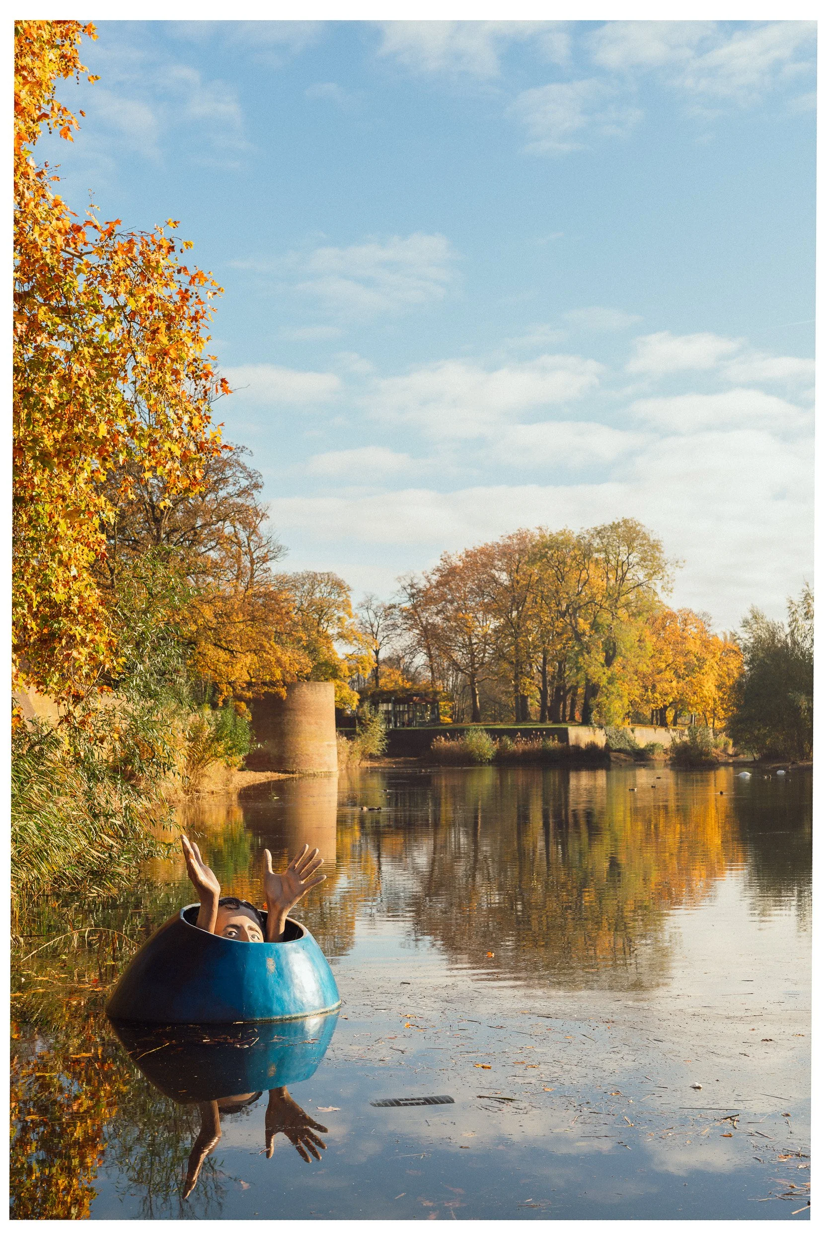 Een persoon met alleen het hoofd en handen zichtbaar, uit een blauwe bootje in een rivier, omgeven door herfstbladeren en bomen, met een blauw hemel in de achtergrond.