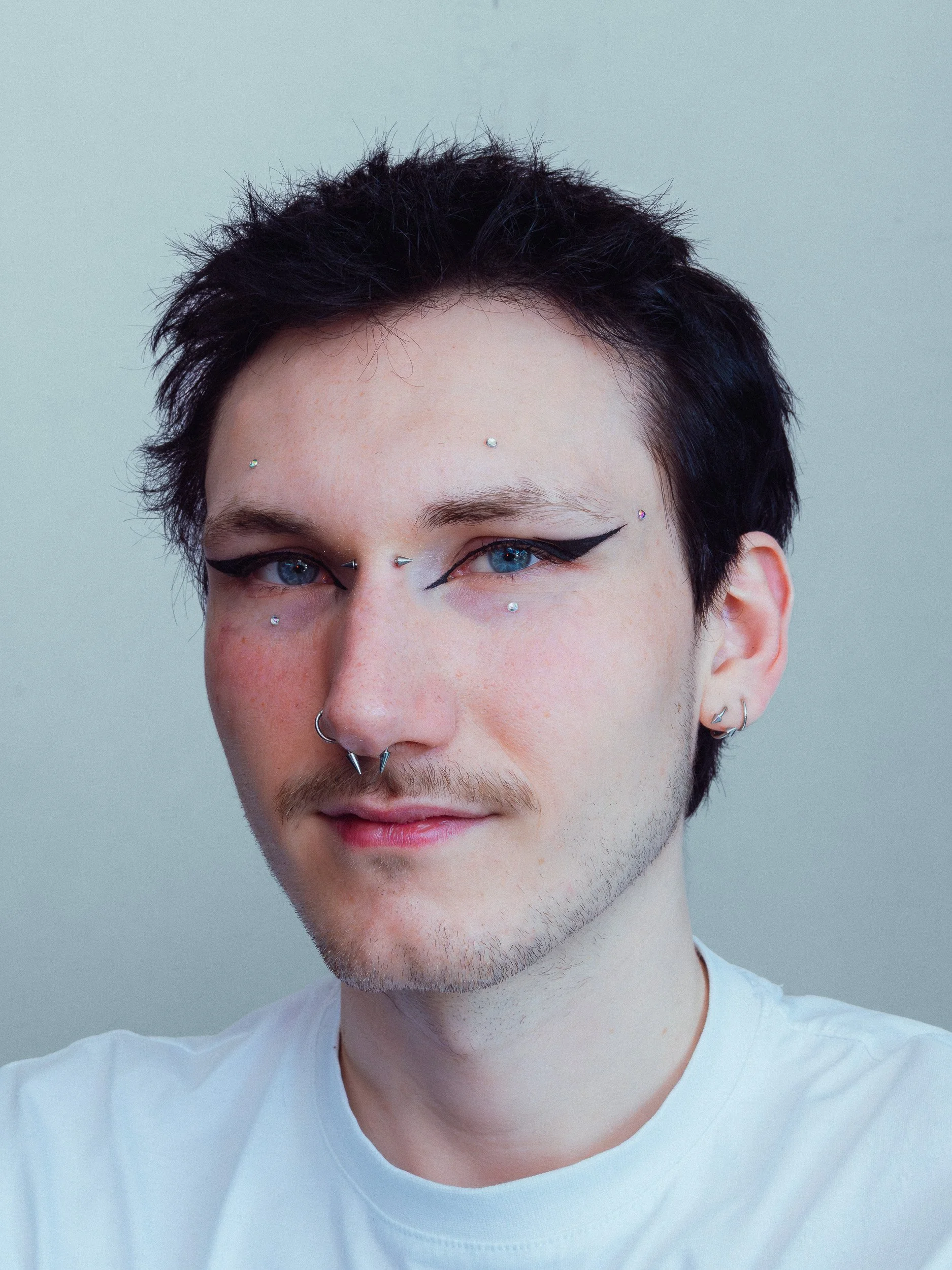 Close-up van een jonge man met spposjes in het gezicht, zwart haar, blauwe ogen en dragen een witte T-shirt, met lichte grijze achtergrond.