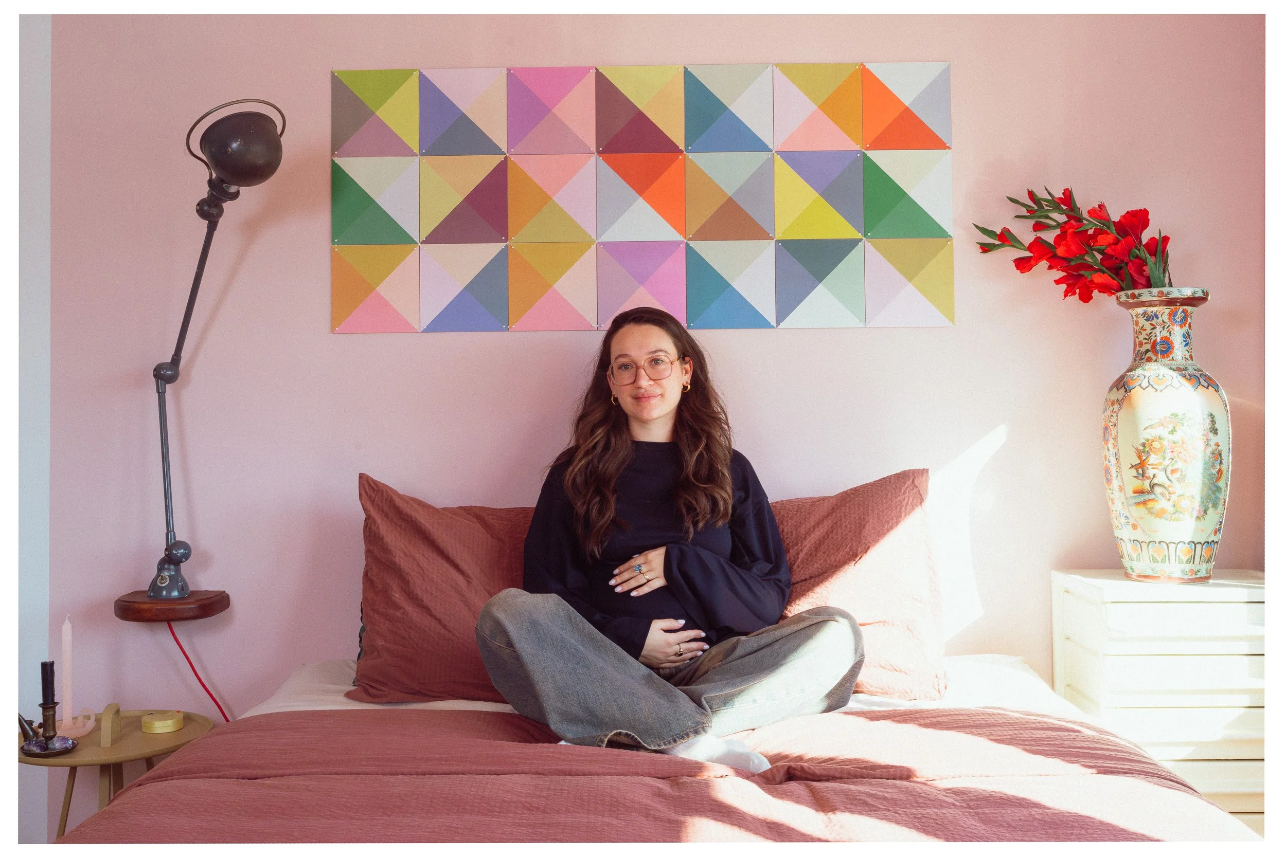 Vrouw met lang bruin haar, bril en ring, zittend op een bed met roze en grijze kleding, in een kamer met een kleurrijk kunstwerk, een grote bloemenvaas met rode bloemen, een leeslamp en een nachtkastje.