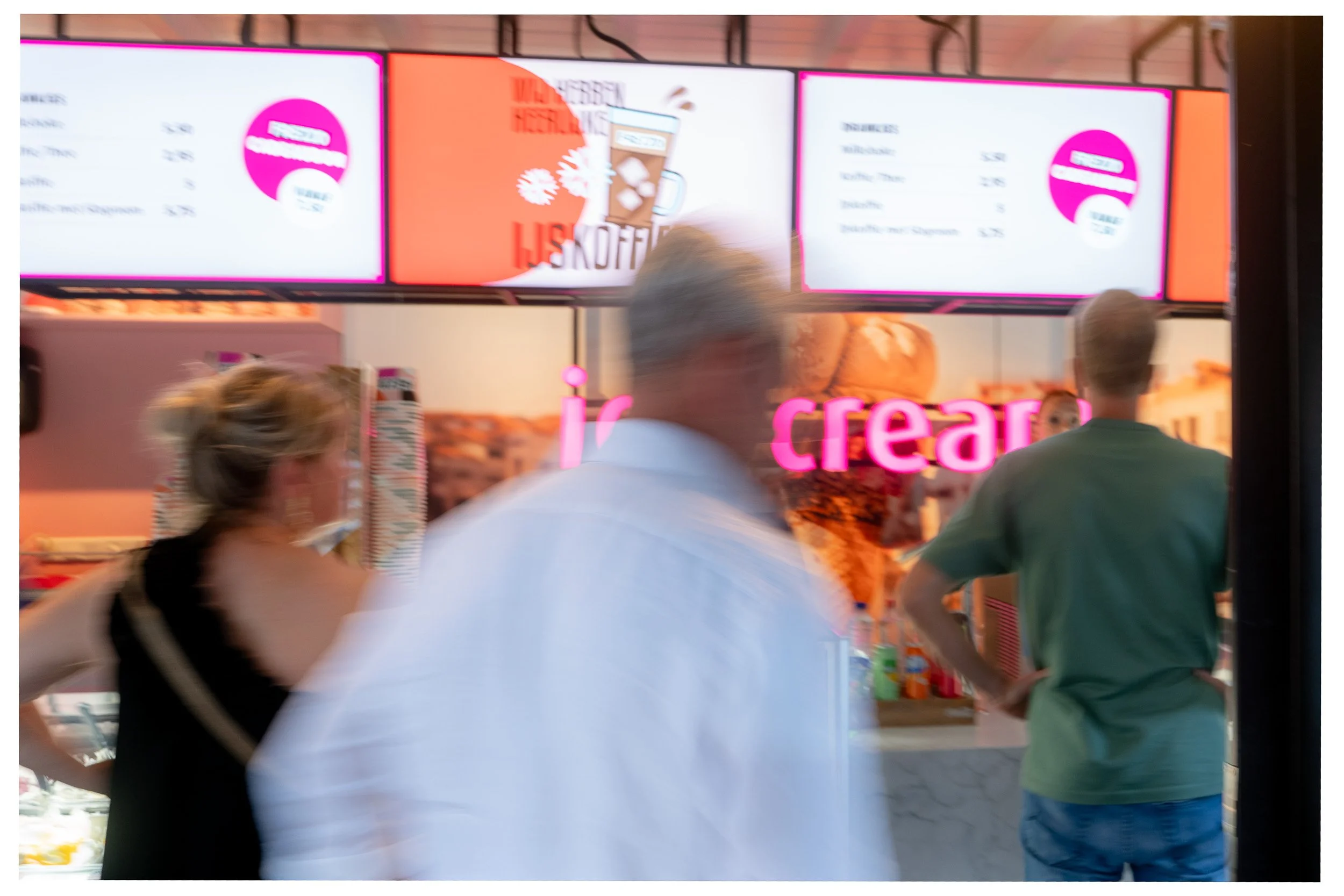 Binnenzicht van een ijs- of gelato winkel met drie mensen die wachten of bestellen, met digitale menu’s boven de deur die prijzen en producten tonen.