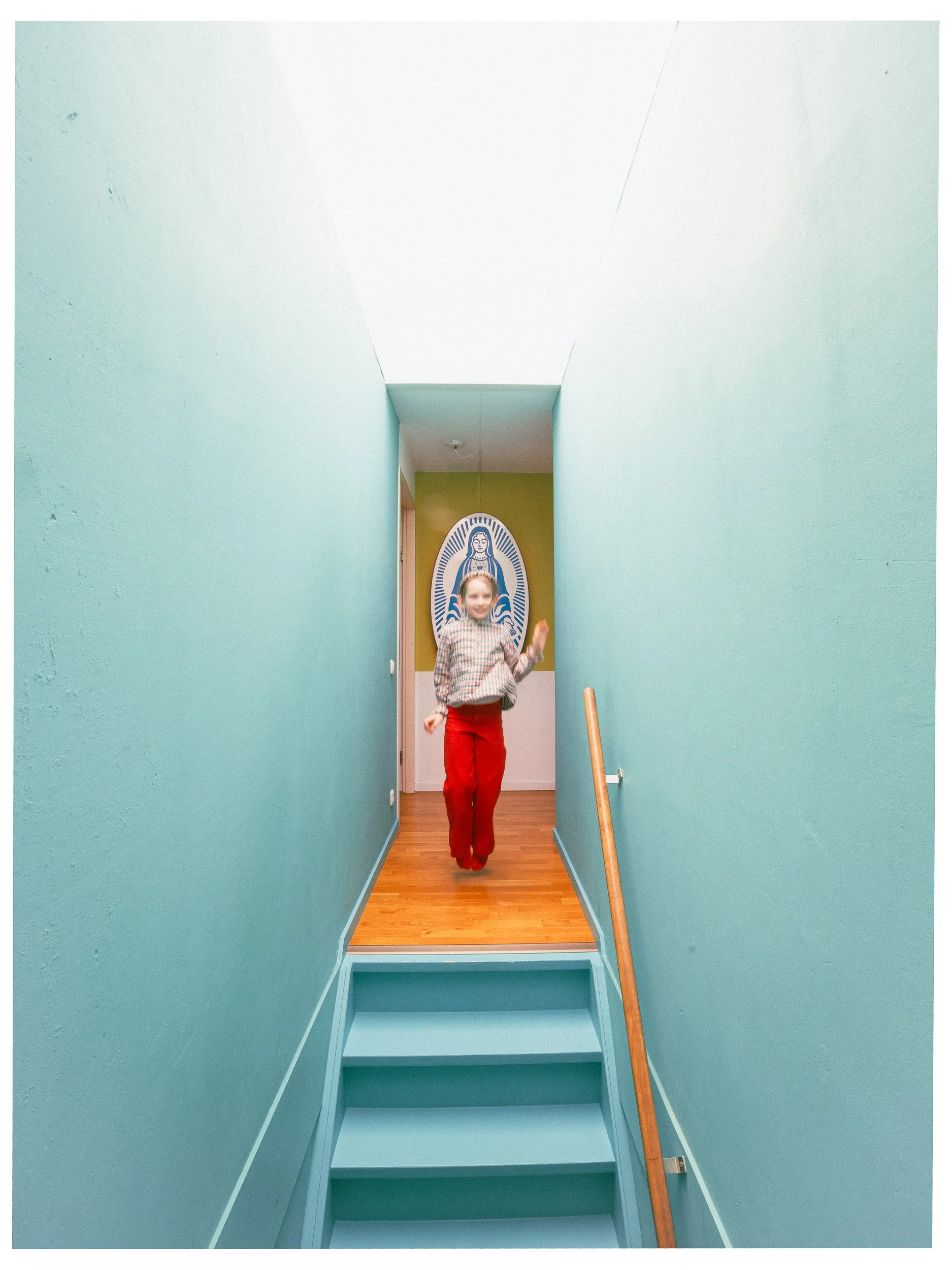 Een kind in een kamer met mintgroene muren en een houten vloer, dat springt op een trap naar de kamer met een afbeelding van de Maagd Maria op de muur.