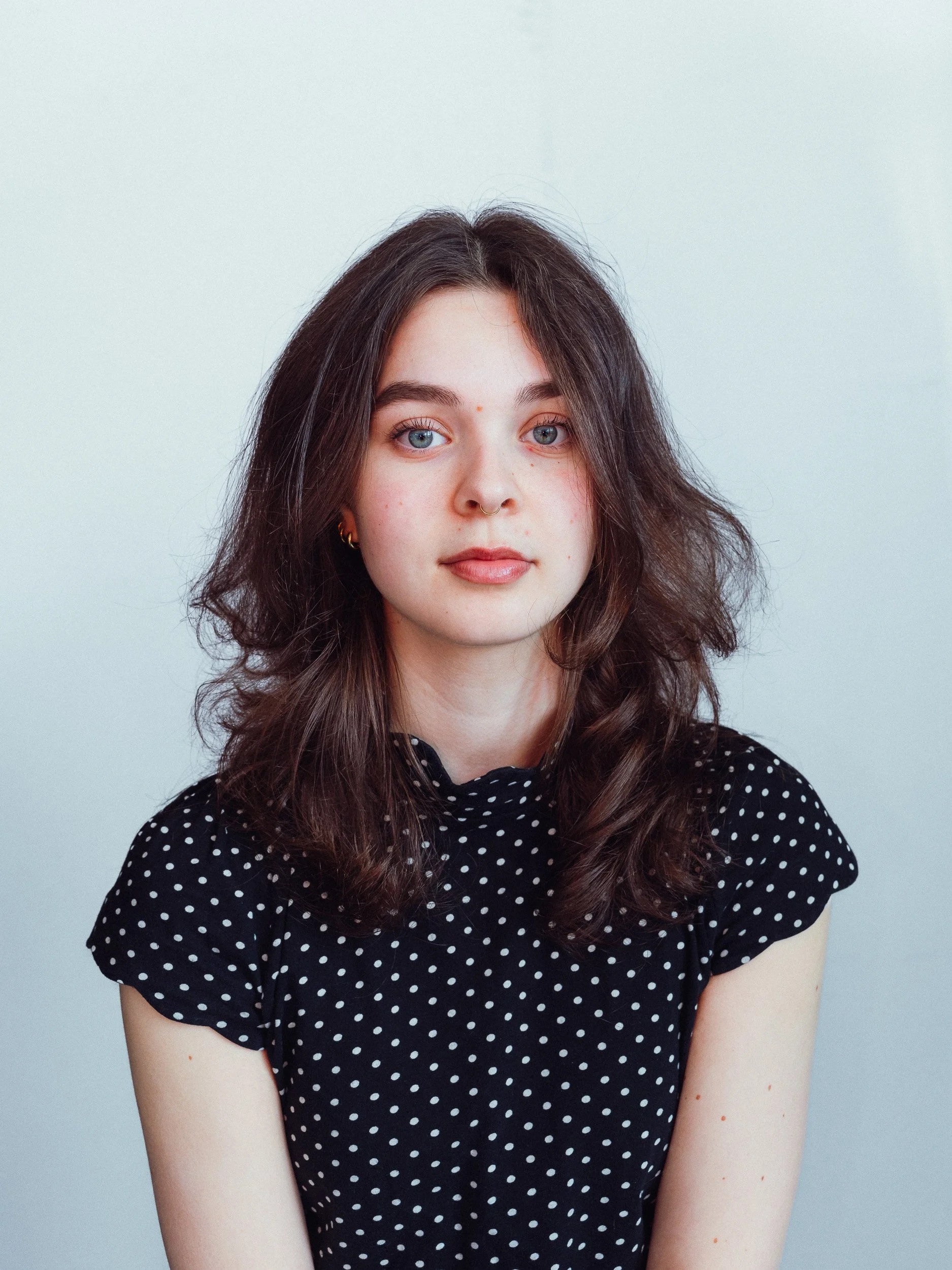 Portret van een jonge vrouw met lang, golvend bruin haar, blauwe ogen, een neusring en oorbellen, gekleed in een zwarte jurk met witte stippen, voor een lichte achtergrond.