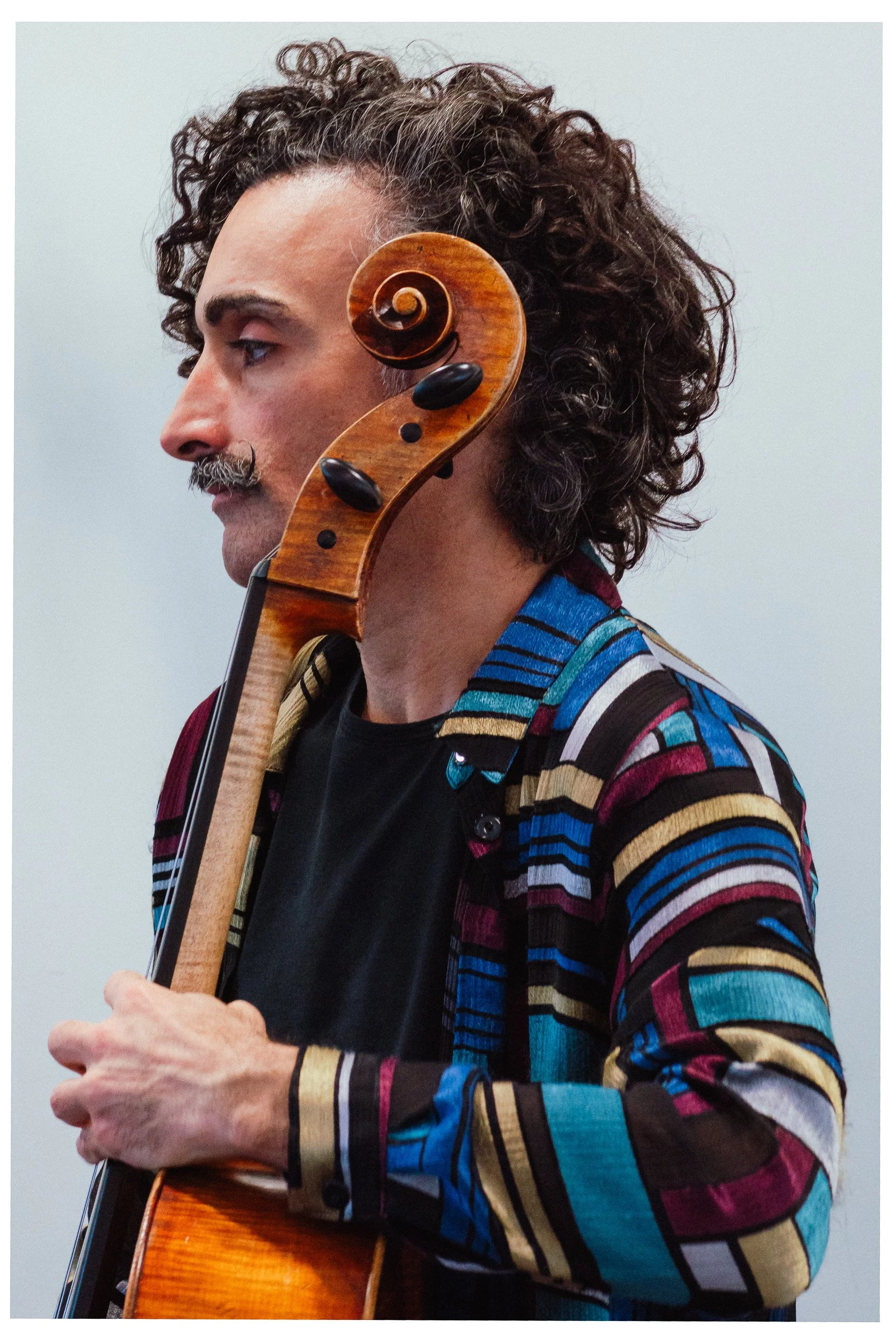 Man met een warrige, gekrulde haardos die een houten cello bespeelt, gekleed in een kleurrijk, gestreept shirt.