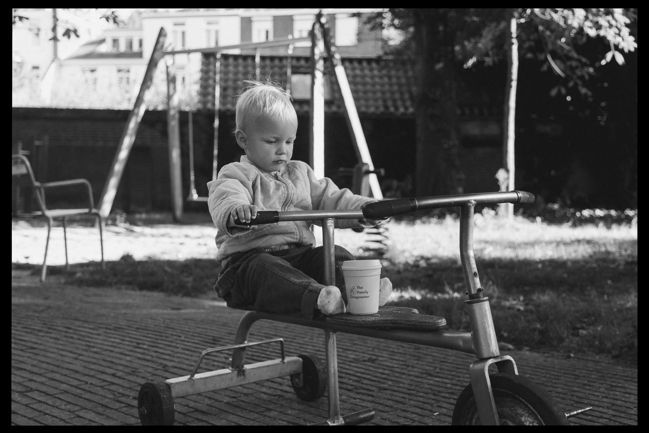een jonge jongen zit op een driewieler in een speeltuin, met een beker op het voorzitje, op de achtergrond een schommel en een boom, in zwart-wit