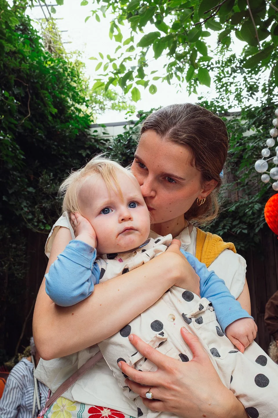 Een vrouw kust een jonge kind op het voorhoofd in een tuin met groene bladeren en een lantaarn in de achtergrond.