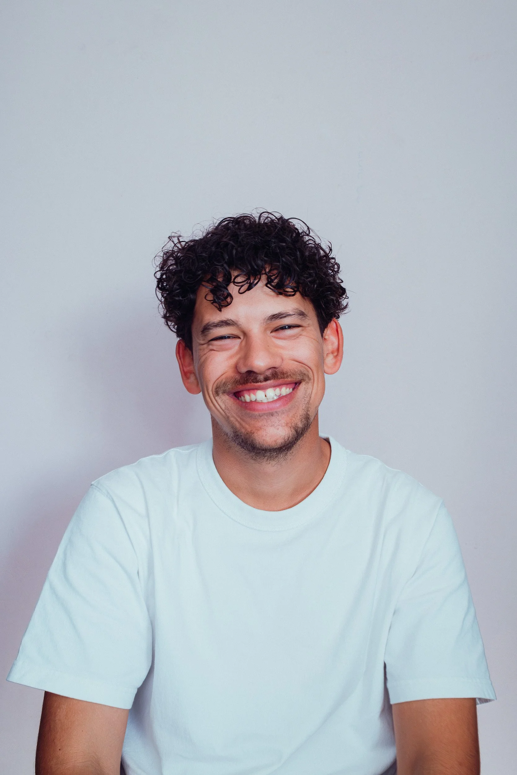 Een lachende man met gekruld haar in een witte T-shirt, lachend naar de camera, tegen een lichtgrijze achtergrond.