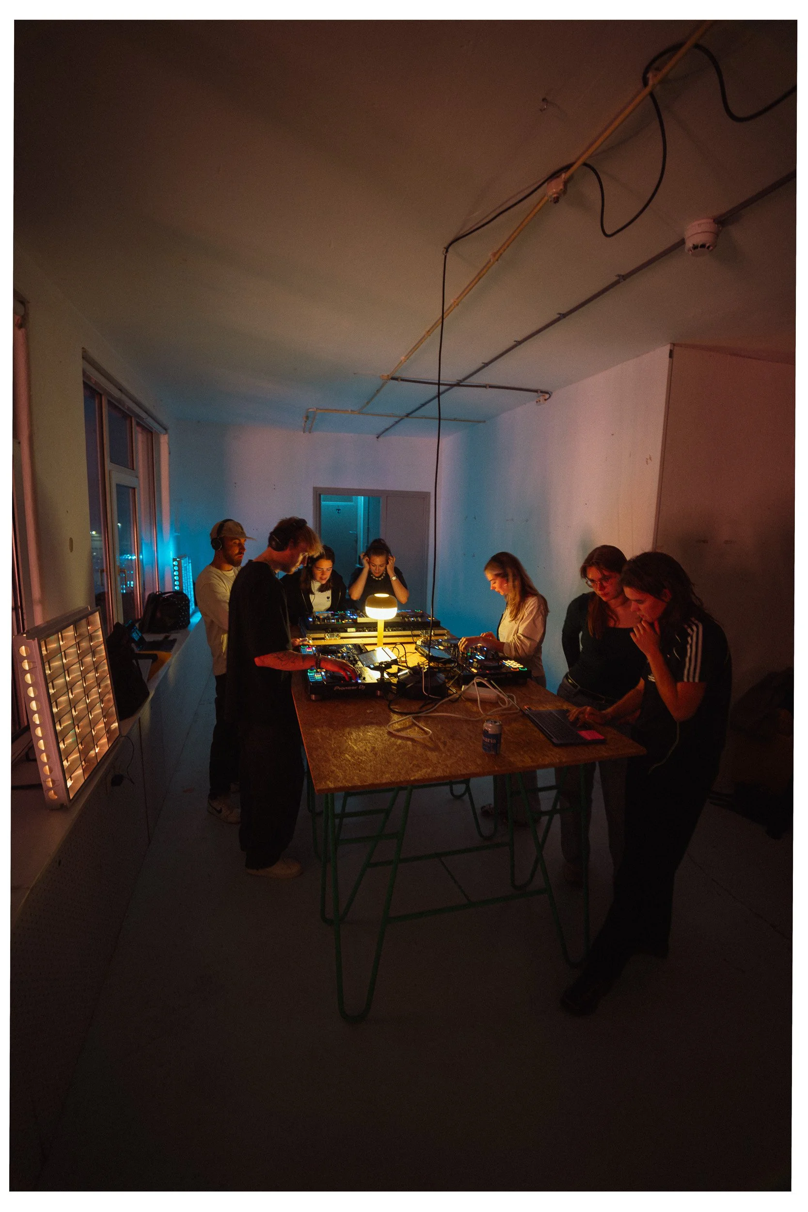 Een groep mensen die in een donkere kamer met sfeervol licht aan het DJ'en is, omringd door DJ-apparatuur en laptops.