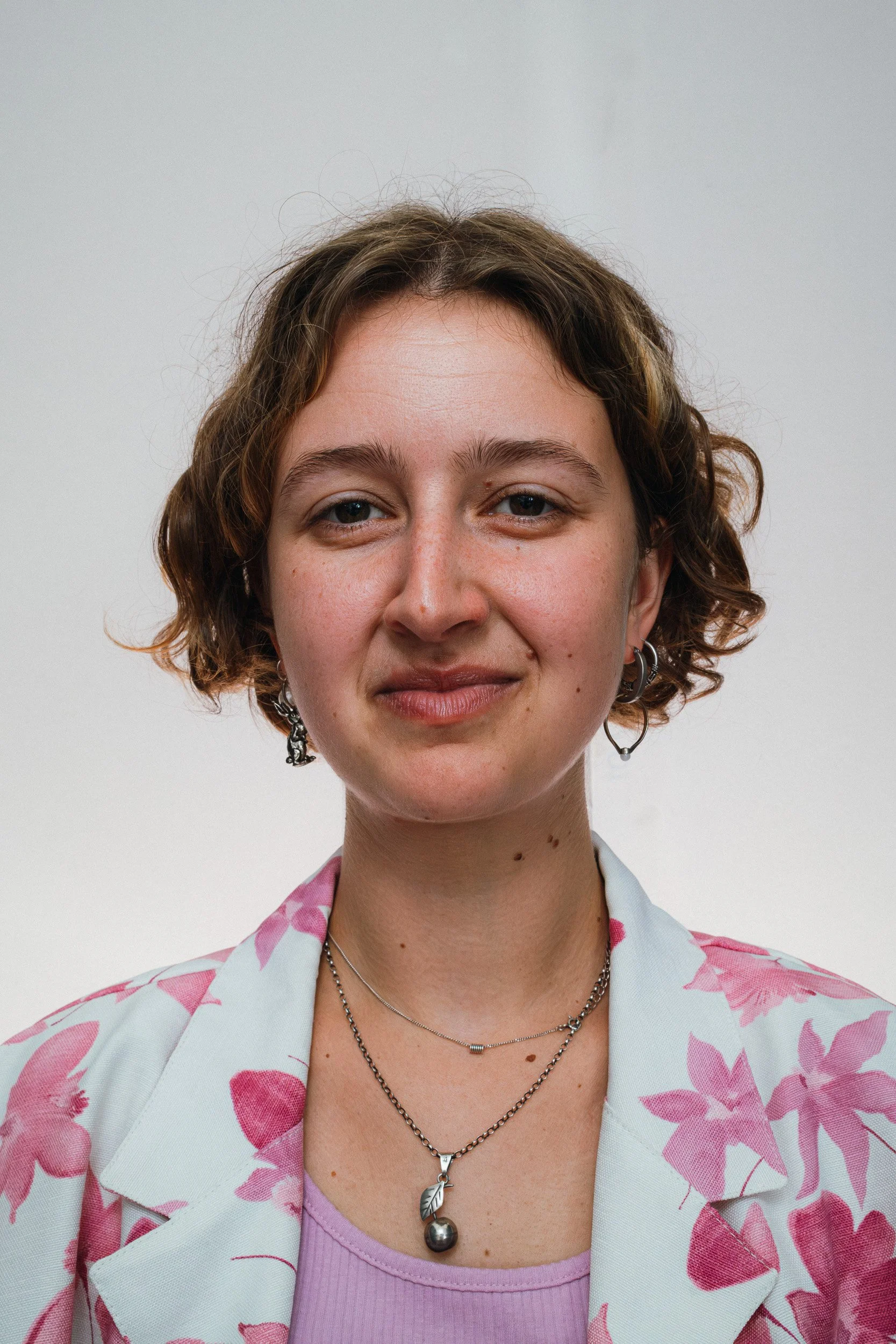 Portret van een jonge vrouw met korte, krullende, bruin haar, die een glimlach heeft en sieraden draagt, gekleed in een witte blazer met roze bloemen en een lila top, voor een effen grijze achtergrond.