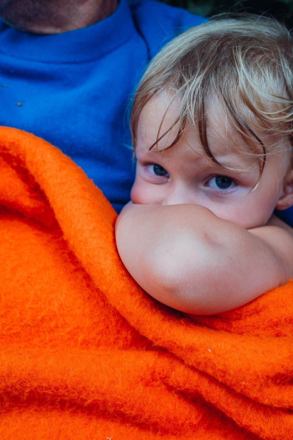 Een jonge jongen met blond haar en blauwe ogen rust met zijn arm over zijn gezicht in een oranje doek, terwijl hij in een blauw shirt zit.