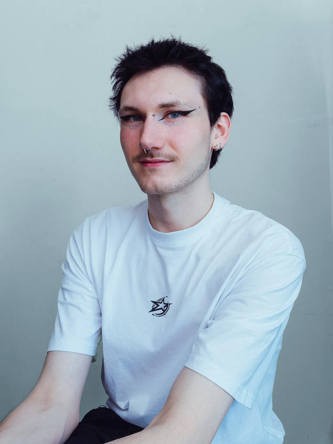 Portret van een jonge man met zwart haar, sieraden en opvallende oogmake-up, zittend voor een effen grijze achtergrond.