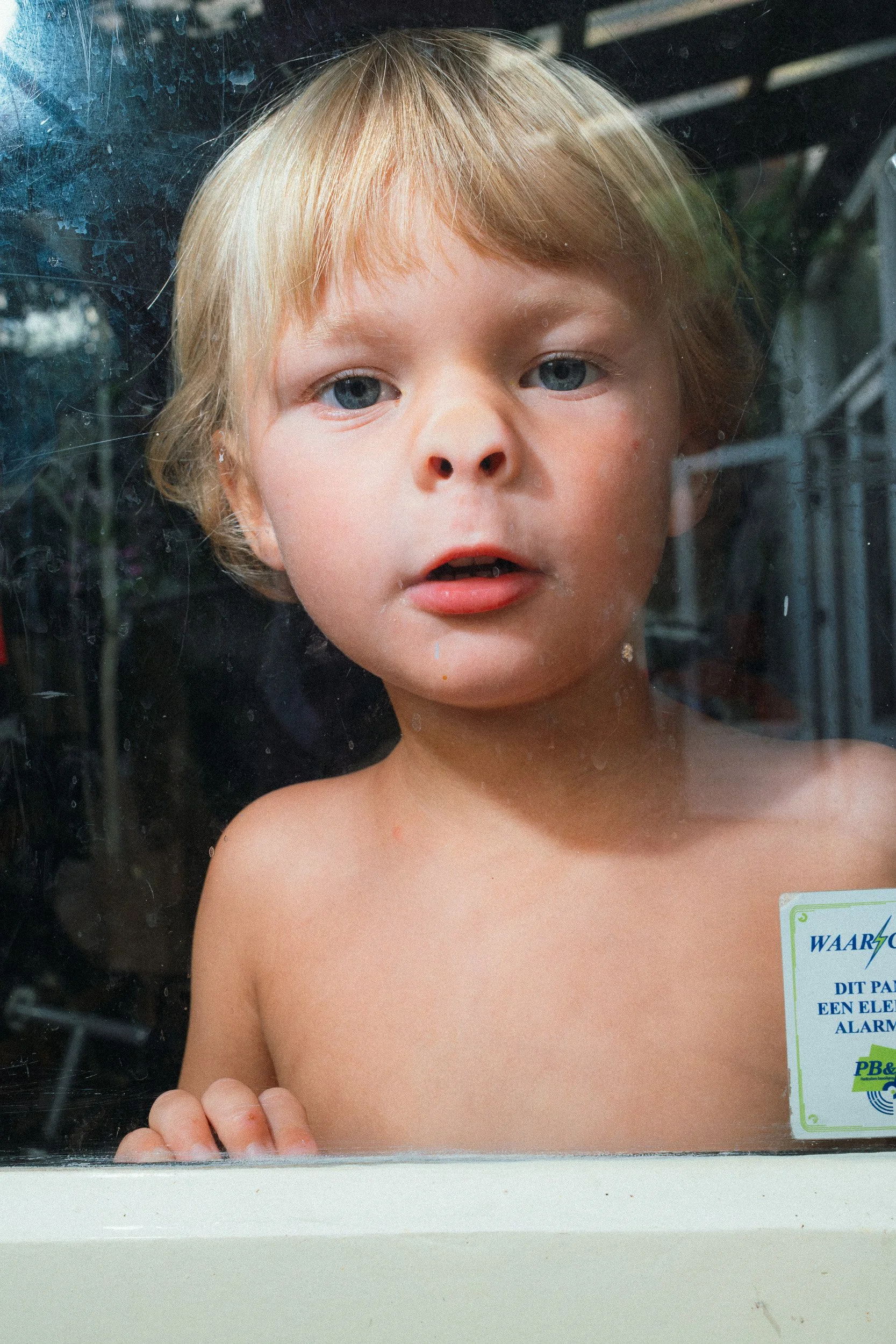 Een jonge jongen zonder shirt met blond haar en blauwe ogen kijkt door een raam met stof en vlekken. Een deel van een bord met tekst is zichtbaar aan de rechterkant.