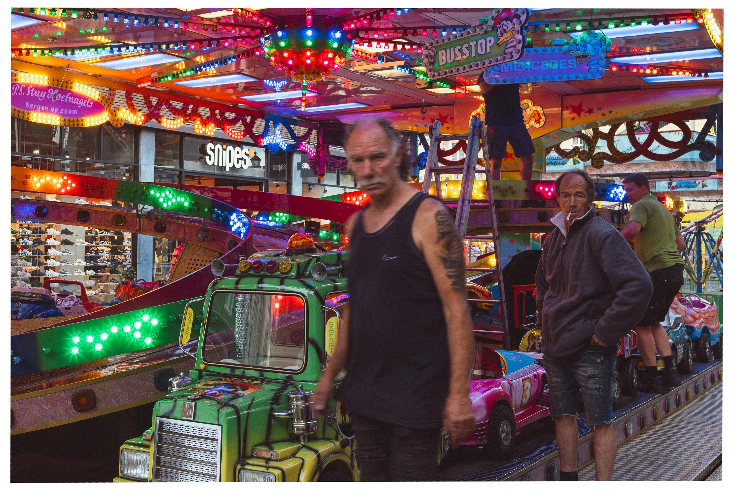 Mensen op een kleurrijk kermisspeelgoed met neonlichten, inclusief een man in een zwart shirt en een man in een grijze jas met een sigaret. Op de achtergrond staan andere mensen, een carrousel en diverse lampjes en decoraties.