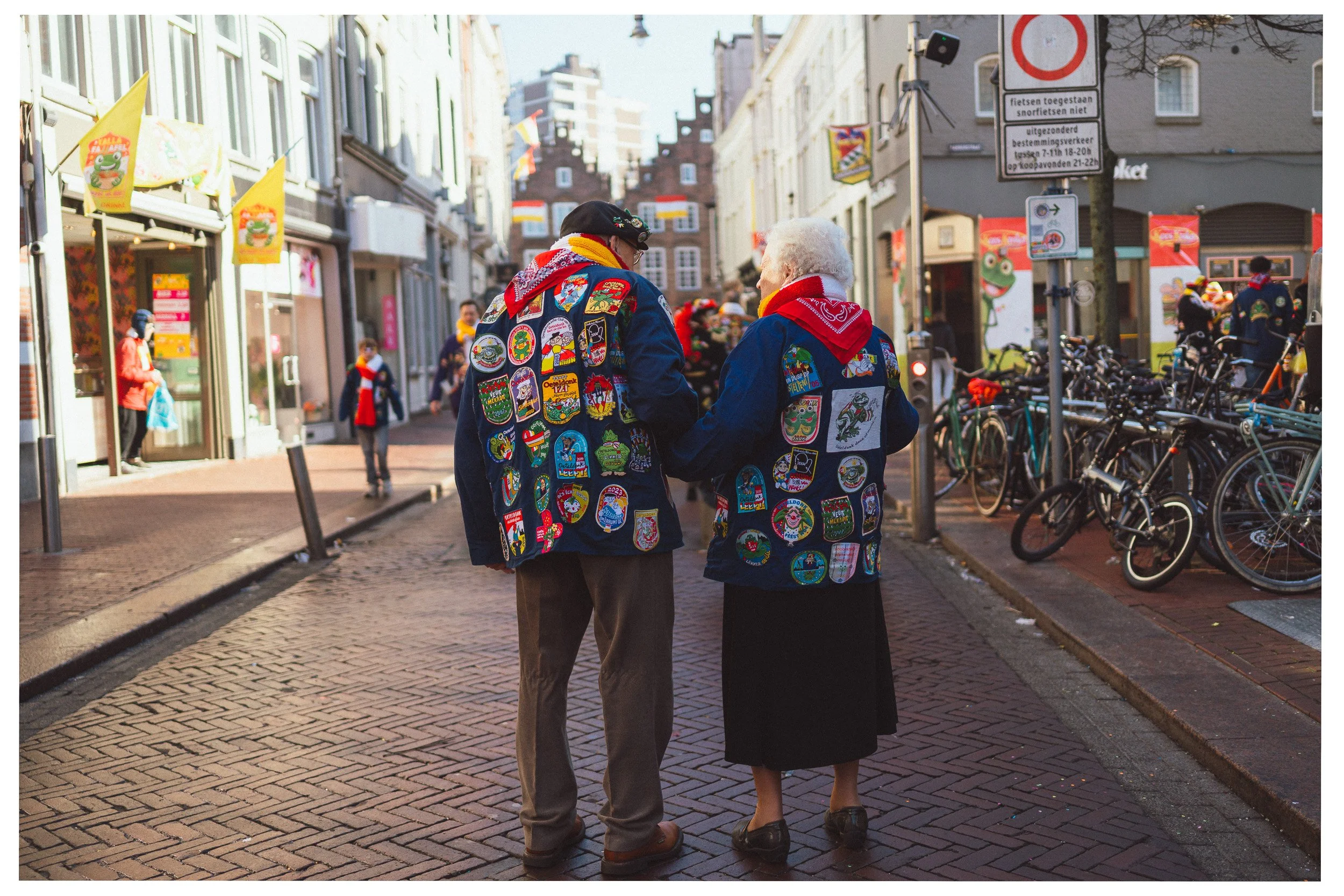 Twee oudere mensen in donkerblauwe jasjes met kleurrijke badges, in gesprek op een straat in een stad, omgeven door fietsen en winkels.