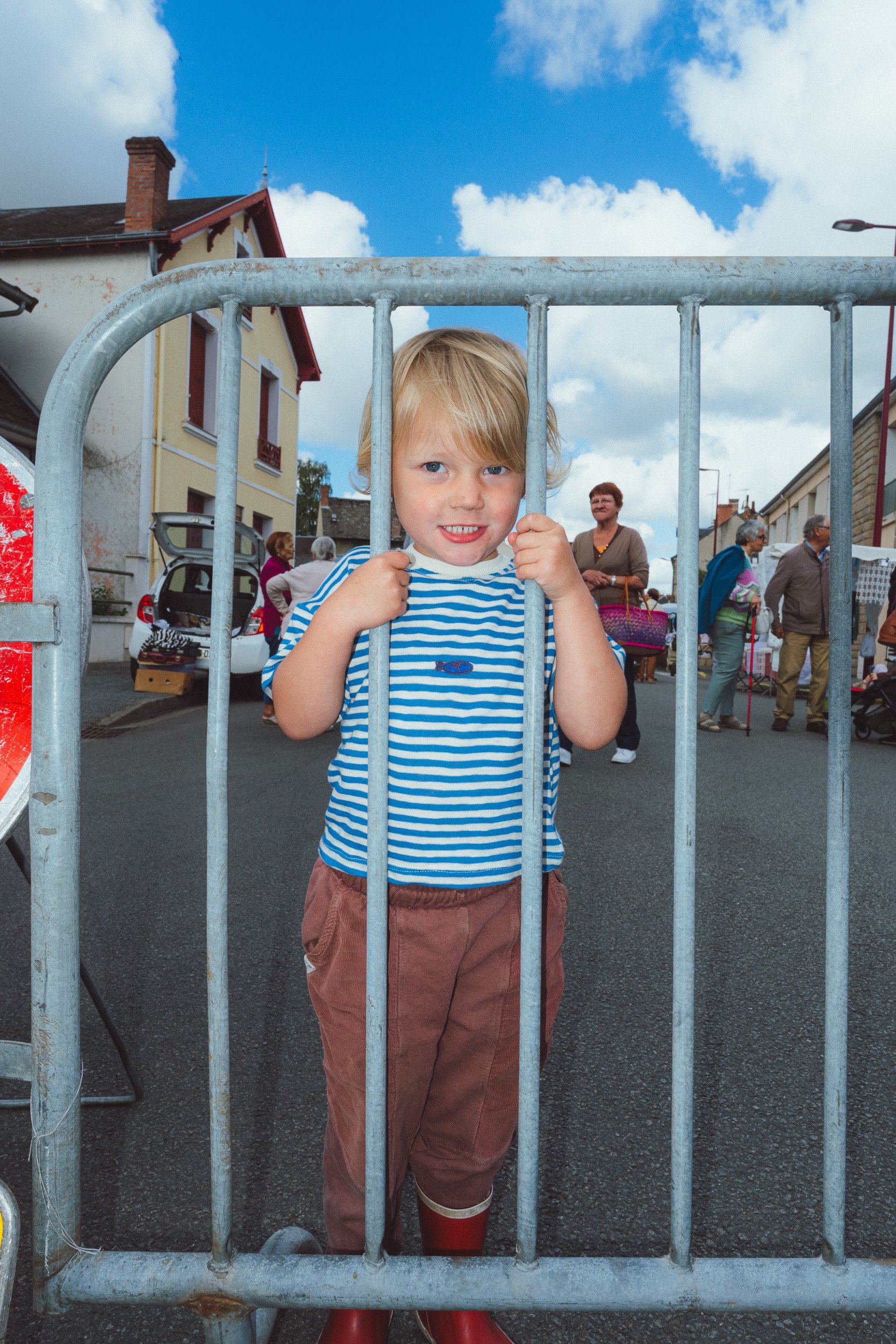 Een jonge jongen met blond haar in een blauwe en witte gestreepte shirt en koraalrode laarzen staat achter een metalen hek, met mensen op de achtergrond en een blauwe lucht met witte wolken.