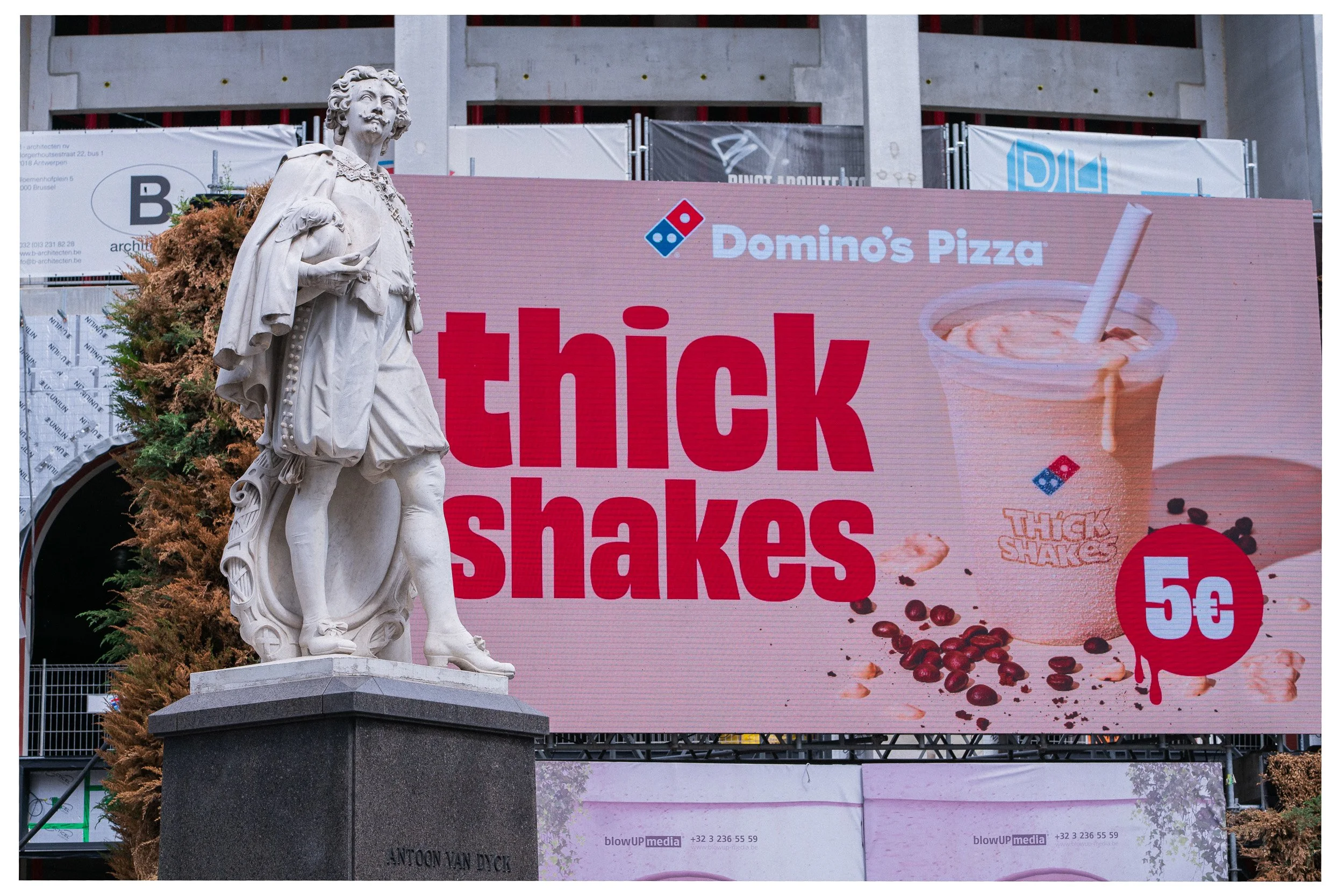 Een standbeeld van een man in historische kleding naast een grote reclamebord voor dikke shakes, met de tekst 'thick shakes' en een afbeelding van een milkshake.